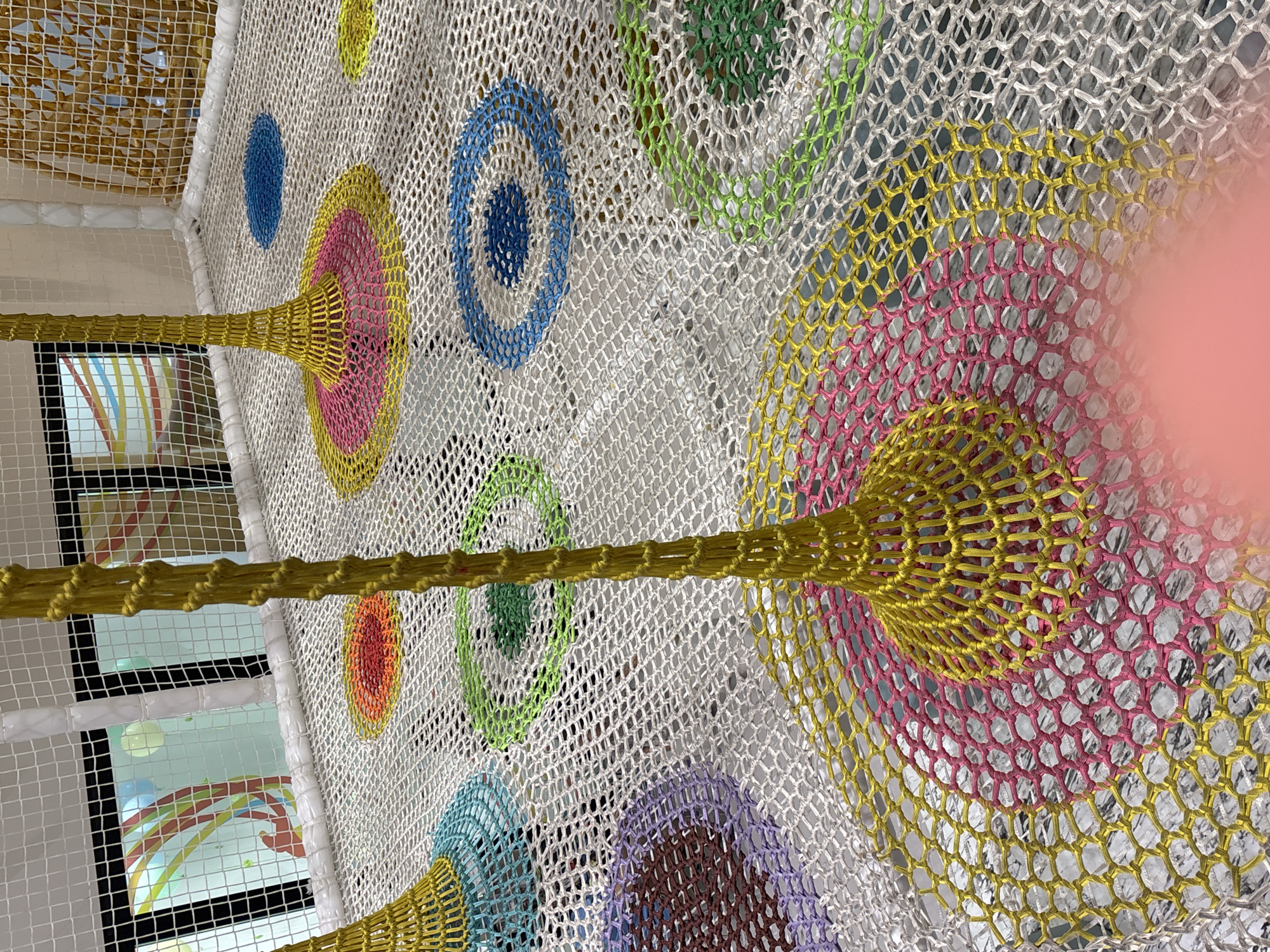 A colorful, netted playground structure is shown from a high angle. The netting is white and forms a mesh floor. Scattered across the floor are circular patches of bright colors: yellow, pink, blue, green, and red. Several yellow, funnel-shaped structures made of the same netting rise from the floor, anchored to the colorful circles. In the background, through a white netted wall, a colorful mural or decoration is visible, possibly on a glass window. The overall impression is one of a playful and vibrant indoor environment.