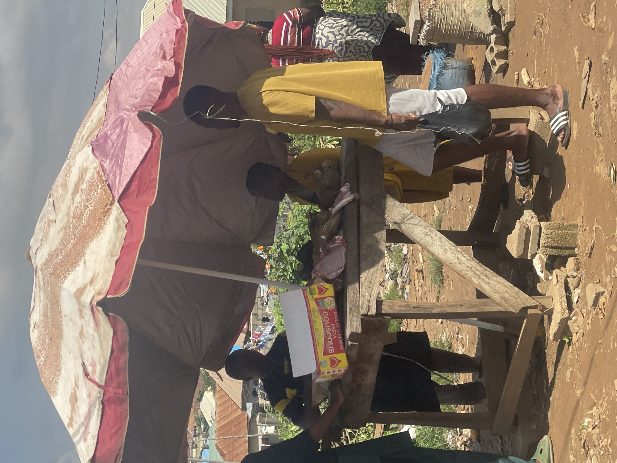 An outdoor market scene is depicted in Kurunduma, Nigeria, beneath a large, worn, multi-colored umbrella providing shade. A wooden table, supported by rough-hewn wooden posts and resting on concrete blocks, displays various items. On the table, a person in a yellow shirt stands to the right, holding a black plastic bag. To their left, another person in a yellow garment faces the table, with a portion of their head visible. In front of them, a person wearing a black shirt with yellow accents is partially visible behind the table, with a box of "GOLDEN PENNY PASTA COUSCOUS" and some raw meat on display. The ground is composed of reddish-brown earth, scattered with debris, small plants, and some discarded materials. In the background, the outlines of buildings and vegetation suggest a semi-urban or peri-urban environment. Another individual, wearing a striped shirt and a patterned skirt, is visible further in the background to the right. The sky is overcast with clouds.