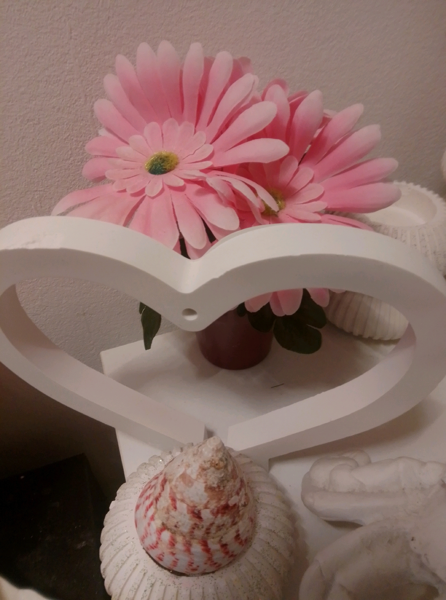 The image depicts a still life arrangement indoors, likely in a home environment. The primary subjects are two artificial pink gerbera daisies with green centers, positioned behind a white heart-shaped frame. The frame is made of a solid material and has a smooth, matte finish. To the right of the flowers and behind the frame, a portion of a white, ridged decorative object, possibly a vase or candle holder, is visible. In the foreground, beneath the heart frame, sits a decorative seashell with red and white spiral patterns, placed on a ridged white base that is sprinkled with glitter. To the right of the seashell, a white, sculptural object, possibly another decorative piece, is partially visible. The background is a textured, pale pink wall. The lighting appears to be artificial and somewhat dim. There is no indication of the city of Wedel, Germany, or any specific location within it.