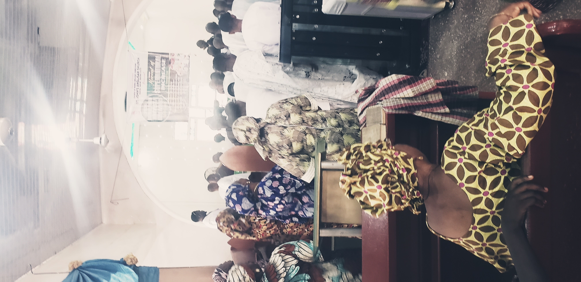 The image shows the interior of St. John's Anglican Church in Kaduna South, Nigeria, during a service. A congregation of people, many wearing traditional attire with vibrant patterns and headwraps, are seated on pews. In the foreground, a woman with a patterned headwrap and dress is seen from behind, resting her arm on the pew. The pews are made of dark wood. Behind the congregants, a banner hangs from the wall, advertising the "Harvest of Supernatural Increase." The room has a tiled ceiling with ceiling fans and fluorescent lighting. A man in a white garment stands facing the congregation in the background. The overall atmosphere appears to be one of communal worship and gathering.