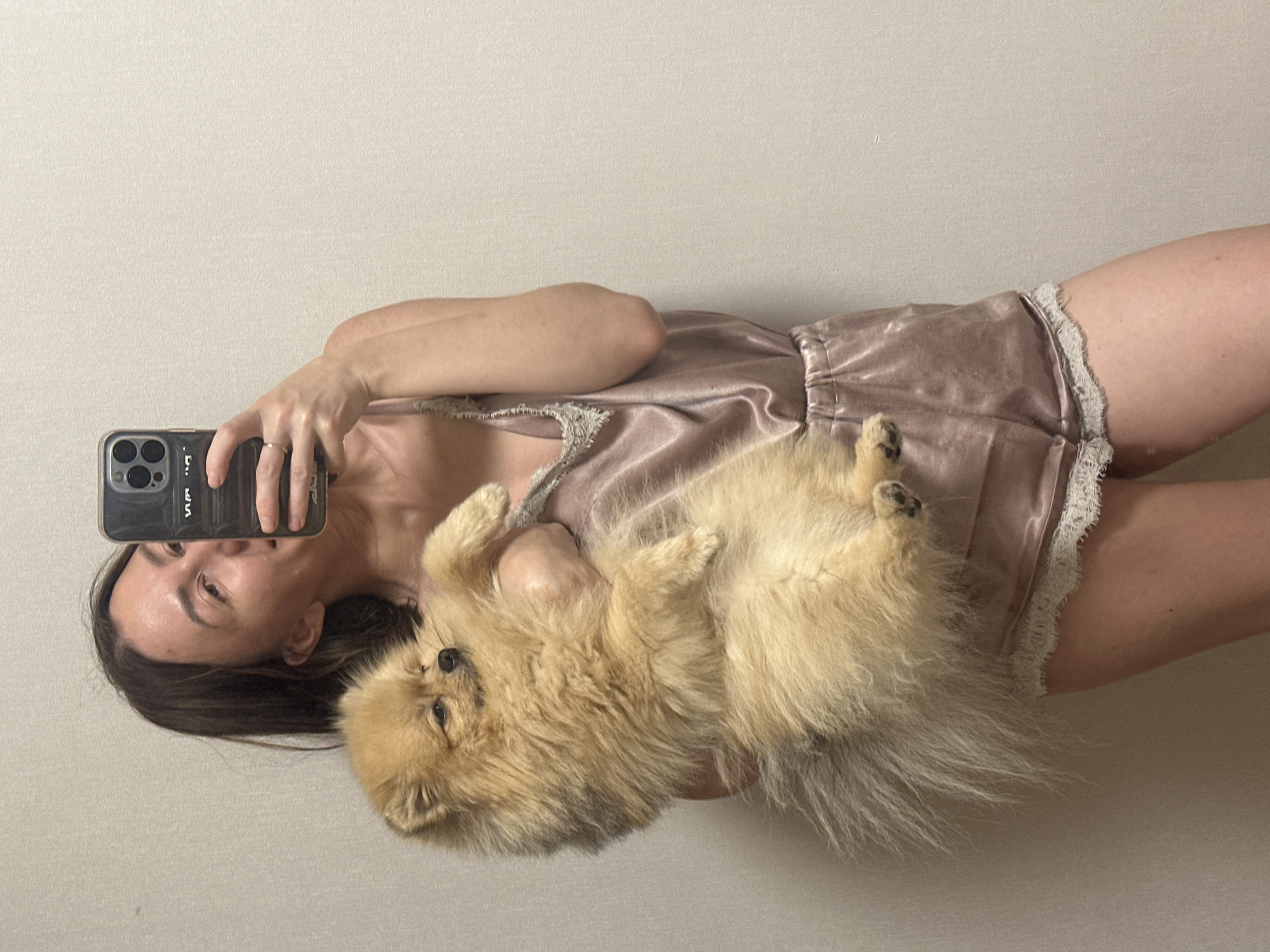A woman stands in front of a plain wall, taking a selfie with her phone. She is holding a fluffy Pomeranian dog, which is nestled in her arms. She is wearing a silky pink camisole with lace trim and matching shorts. The dog is facing away from the camera, with its eyes closed. The background is a neutral, light-colored wall. No specific location in Votkinsk, Russia, can be determined from the image.