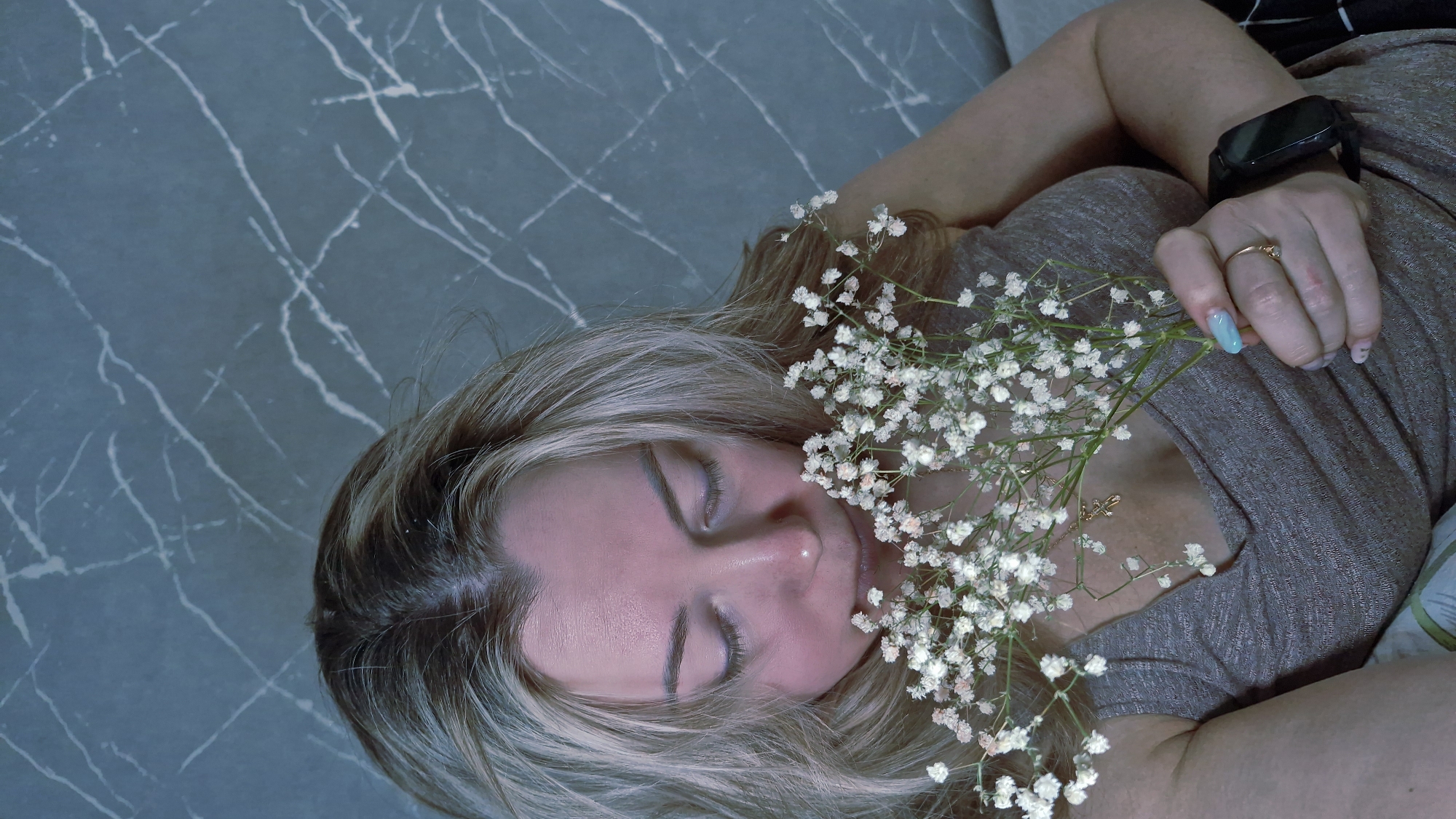 This image captures a peaceful scene of a woman resting indoors. The main subject is a woman, likely Caucasian, with blonde or light brown hair, lying down with her eyes closed. Her expression appears serene and calm, suggesting she might be asleep or deeply relaxed.
She is dressed in a simple grey or light brown short-sleeved top. A delicate sprig of small white flowers, characteristic of Baby's Breath (Gypsophila), rests on her chest and close to her face, adding a touch of softness and natural beauty to the composition.
Notable accessories include a black smartwatch with a rectangular screen on her left wrist and a plain gold ring on the ring finger of her right hand. Her fingernails on her right hand are painted with a light blue polish. A subtle gold necklace with a small pendant is also visible on her décolletage, partially hidden by the flowers.
The woman is positioned diagonally against a background that features a grey-blue surface with distinct white, marble-like patterns or veins. This surface could be a bedspread, blanket, or flooring, indicating an indoor setting. The lighting is soft and even, contributing to the tranquil atmosphere. No specific time of day or outdoor weather conditions are discernible. There is no visible text in the image. The scene is situated in Otchor, Russia.