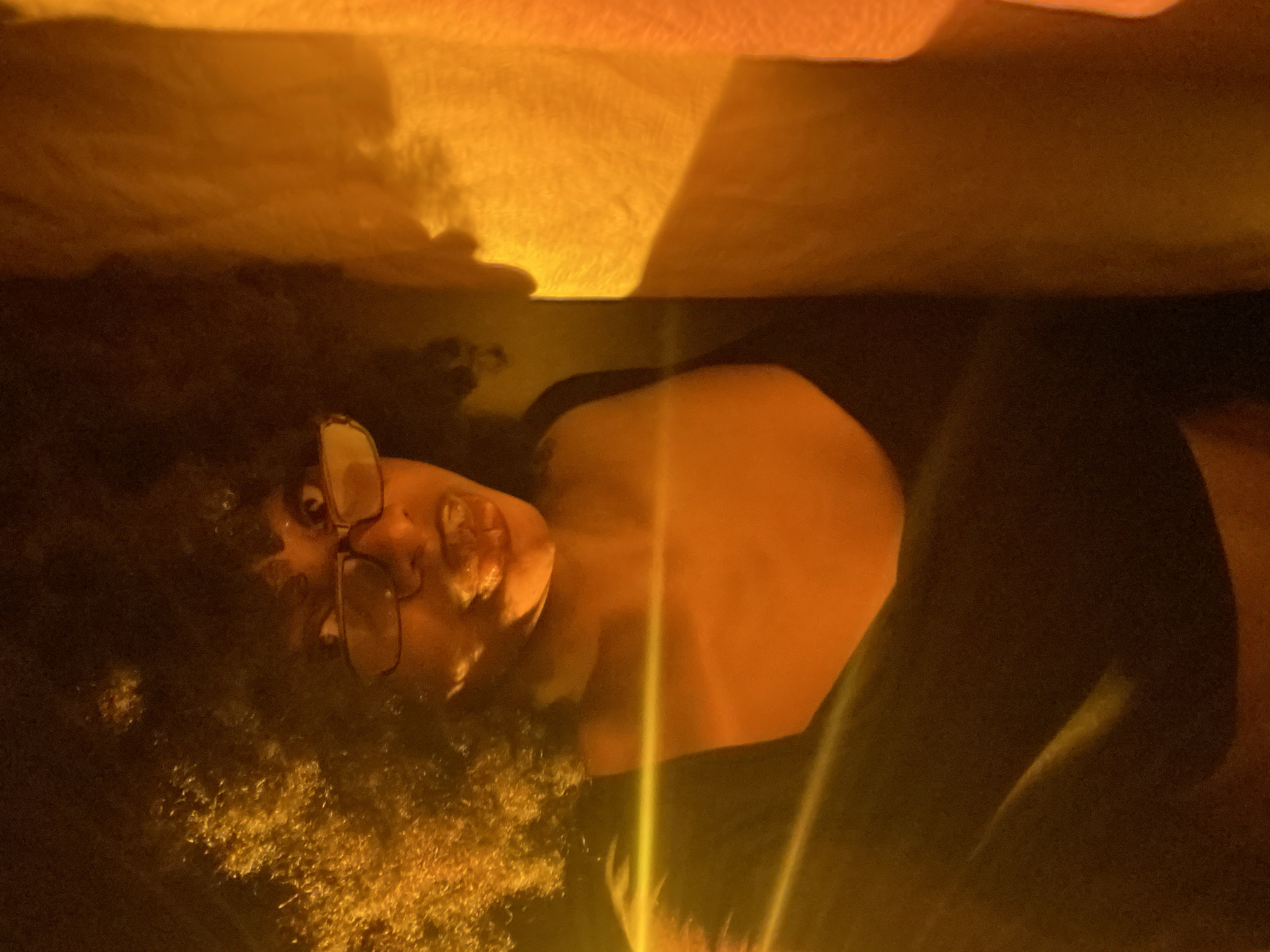 A person with curly hair and glasses is lying down in a dimly lit environment. The person is partially under a yellow cloth or blanket. The scene is illuminated with a warm, yellow light source. The location in the city of Iranla, Nigeria is not discernible from the image.