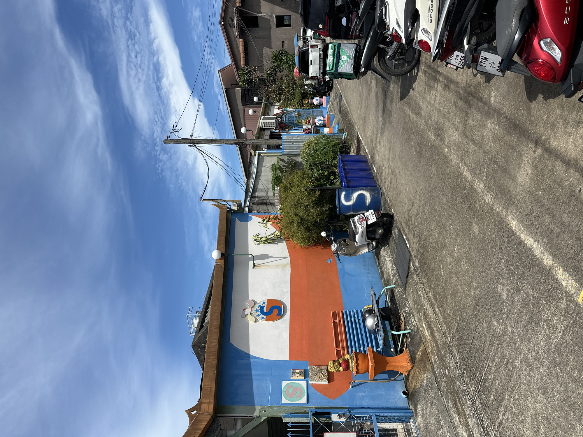 An eye-level, wide shot shows a narrow, paved alleyway in an urban area, possibly in Thailand. The alley is lined with buildings on the left and right, with a blue-sided building prominently featured on the left. This building is decorated with a white section containing a large orange 'S' logo with blue stars and what appears to be a cartoon rabbit. Below this, an orange stripe runs across the blue wall. To the left of the building is a blue metal gate. In front of the blue building, a blue bench with two helmets on it sits next to an orange pedestal topped with a decorative lion statue.
On the right side of the alley, several motorcycles and a white SUV are parked. Some of the motorcycles have delivery bags attached. Further down the alley, more buildings are visible, including a multi-story structure. The sky above is blue with scattered white clouds. The overall scene depicts a typical street or alley in a developed area, with elements of residential and commercial activity. There are no people visible in the image.