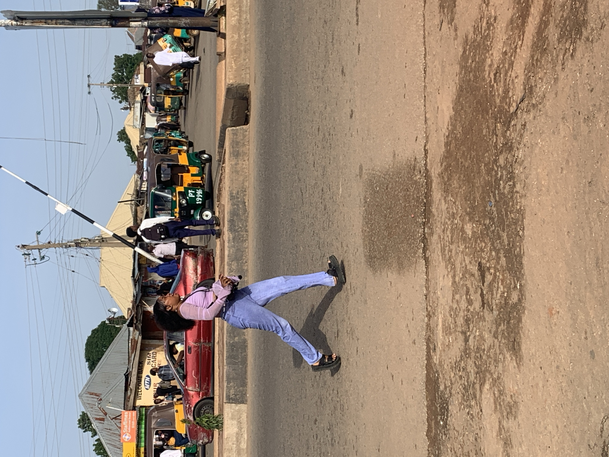 In Gura Top, Nigeria, a young woman with dark, curly hair and a light purple, long-sleeved crop top and light blue jeans walks across a paved road. She is wearing black sandals and carrying a dark bag. The road is in front of a line of buildings, some with corrugated metal roofs. A red, older model sedan is parked in front of one of the buildings, with its driver's side door open. Various auto-rickshaws, some green and yellow, are parked or moving on the road. A white barrier arm, likely for traffic control, is visible in the upper part of the image. The environment appears to be a busy urban area with a clear sky and sunlight casting shadows.