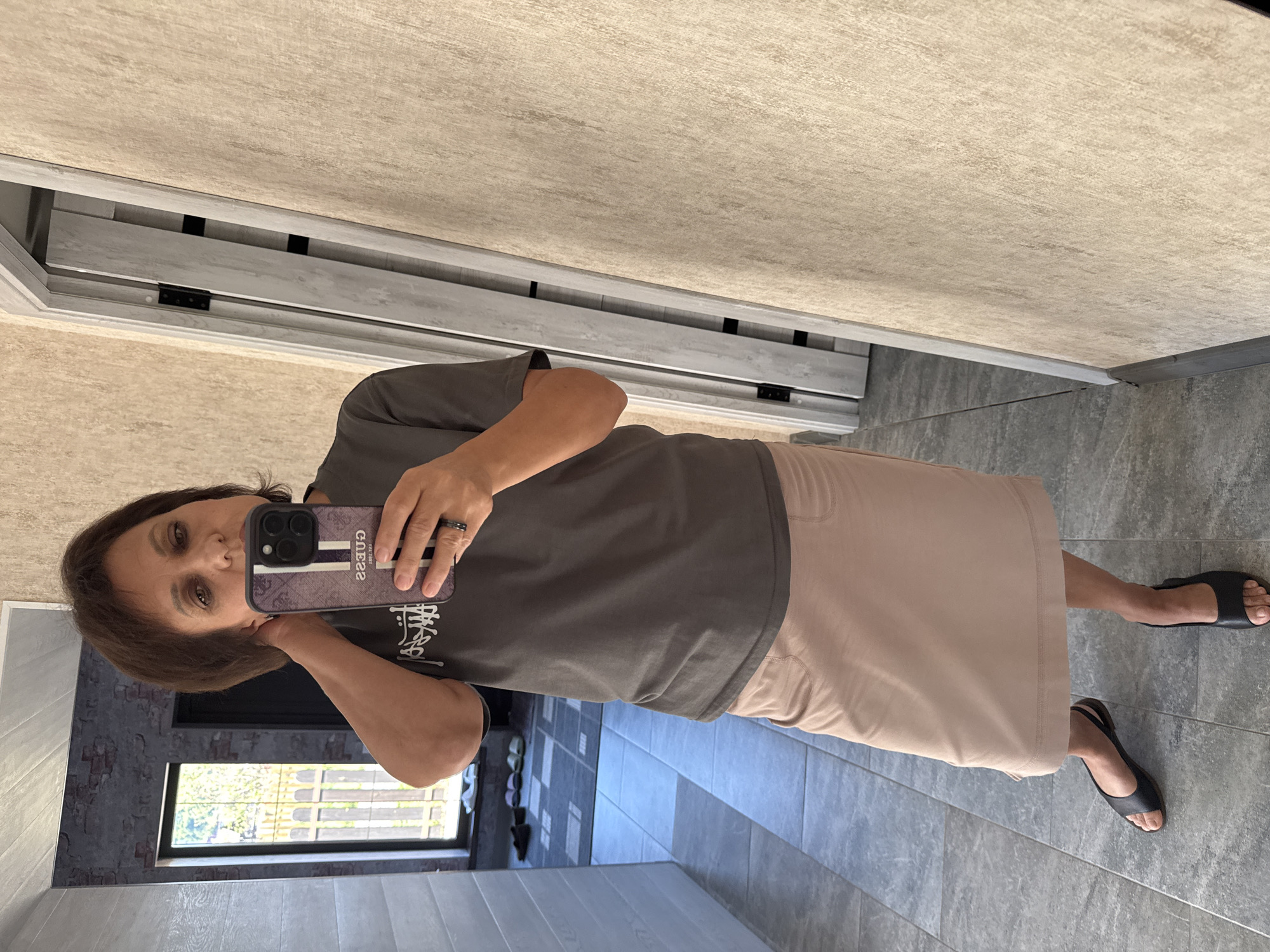 A woman is taking a selfie in a hallway. She is wearing a dark gray t-shirt and a light beige skirt. She has on black slingback sandals. The hallway has gray tiled floors and a light beige textured wall. To her left, there is a mirror reflecting a window with a fence outside and what appears to be a brick-patterned wallpaper. To her right, there is a light-colored door that is partially open, revealing more of the door. The location is identified as Soloneshnoye, Russia.