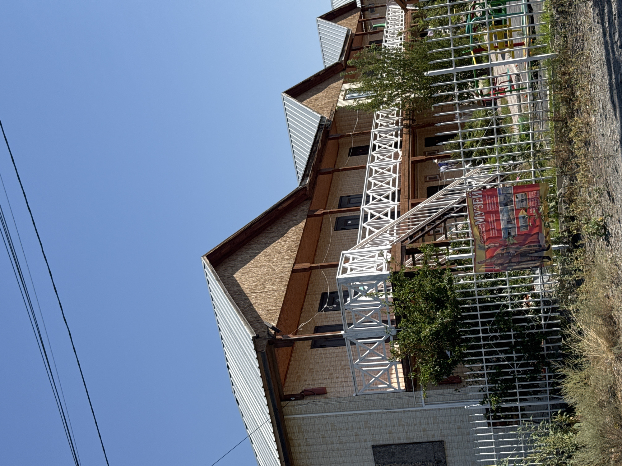 The image depicts a row of multi-story buildings with metal roofs, wooden accents, and balconies, possibly serving as accommodation or a resort. A white metal fence encloses the property, with a playground visible in the background. A red poster with text in Cyrillic, including the word "МУЗЕЙ" (Museum), is attached to the fence. Vegetation, including trees and bushes, is present both inside and outside the fence. The sky is clear and blue. The scene is set in Zharbulak, Kazakhstan.