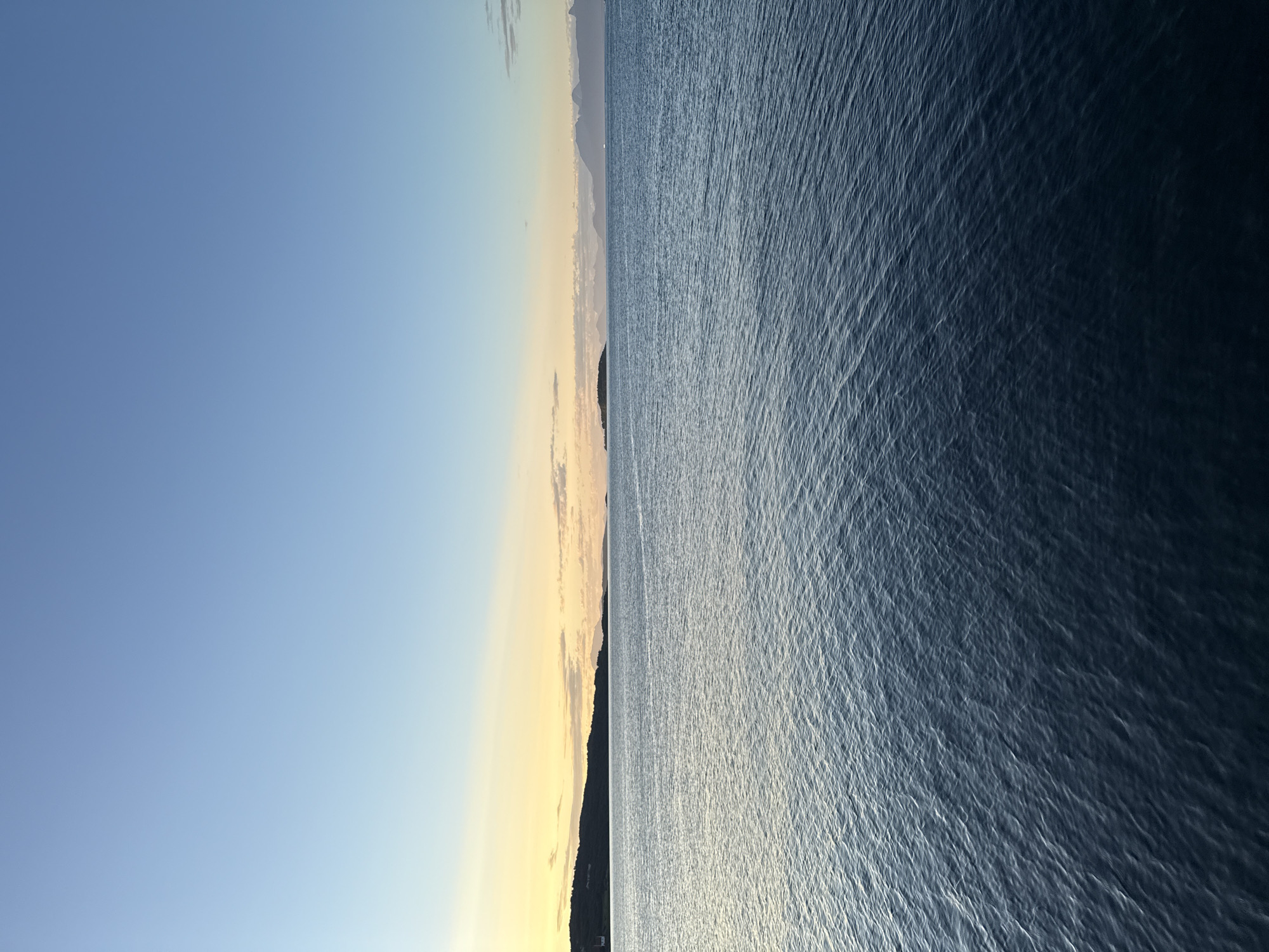 A tranquil seascape unfolds under a gradient sky of soft blues and yellows, indicative of either sunrise or sunset. The vast expanse of the Norwegian Sea dominates the foreground, its surface rippling with gentle waves that reflect the ambient light. In the mid-ground, a distant, dark, and tree-covered island or peninsula stretches horizontally across the horizon, its silhouette contrasting with the brighter sky. Further in the distance, a chain of hazy, mountainous islands creates a majestic backdrop, their peaks softened by atmospheric perspective. The scene evokes a sense of calm and natural beauty. Based on the geography, this view likely looks out towards the west from the city of Harstad, Norway, with the islands of Andøya and perhaps parts of Vesterålen visible in the far distance.