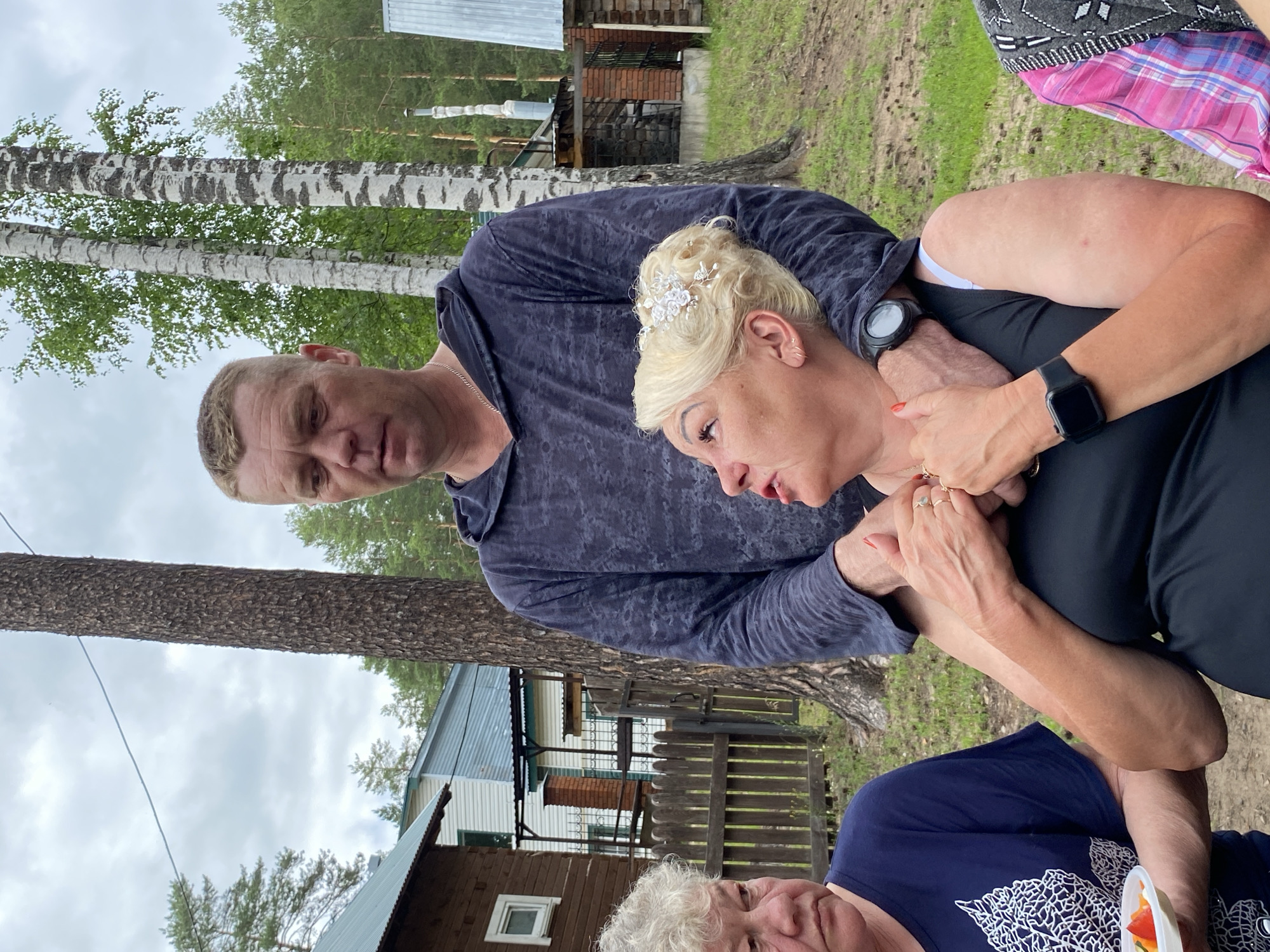 The image captures a candid moment of three individuals outdoors, likely in a rural or semi-rural setting given the presence of trees, a wooden fence, and rustic buildings. Two adults, a man and a woman, are prominently featured in the foreground, embracing. The man, with short blonde hair, wears a dark grey hoodie. The woman, with blonde curly hair adorned with a white floral accessory, is looking down and to the side, with her hand resting on the man's arm. She is wearing a black top and a watch on her left wrist. Her other hand is clasped with the man's. Their embrace suggests affection or comfort.
Further in the background, a third person, an older individual with grey hair, is partially visible, wearing a dark blue top and holding a small container with some food, possibly fruit. The scene appears to be during daylight, with overcast skies contributing to soft, diffused lighting. The background reveals birch trees, a wooden fence, and glimpses of buildings, one of which seems to be made of wood with a metal roof. Another building has a brick chimney. The overall atmosphere is relaxed and informal, suggesting a casual gathering or outing. The location is identified as Noviy, Russia. No visible text is present in the image.