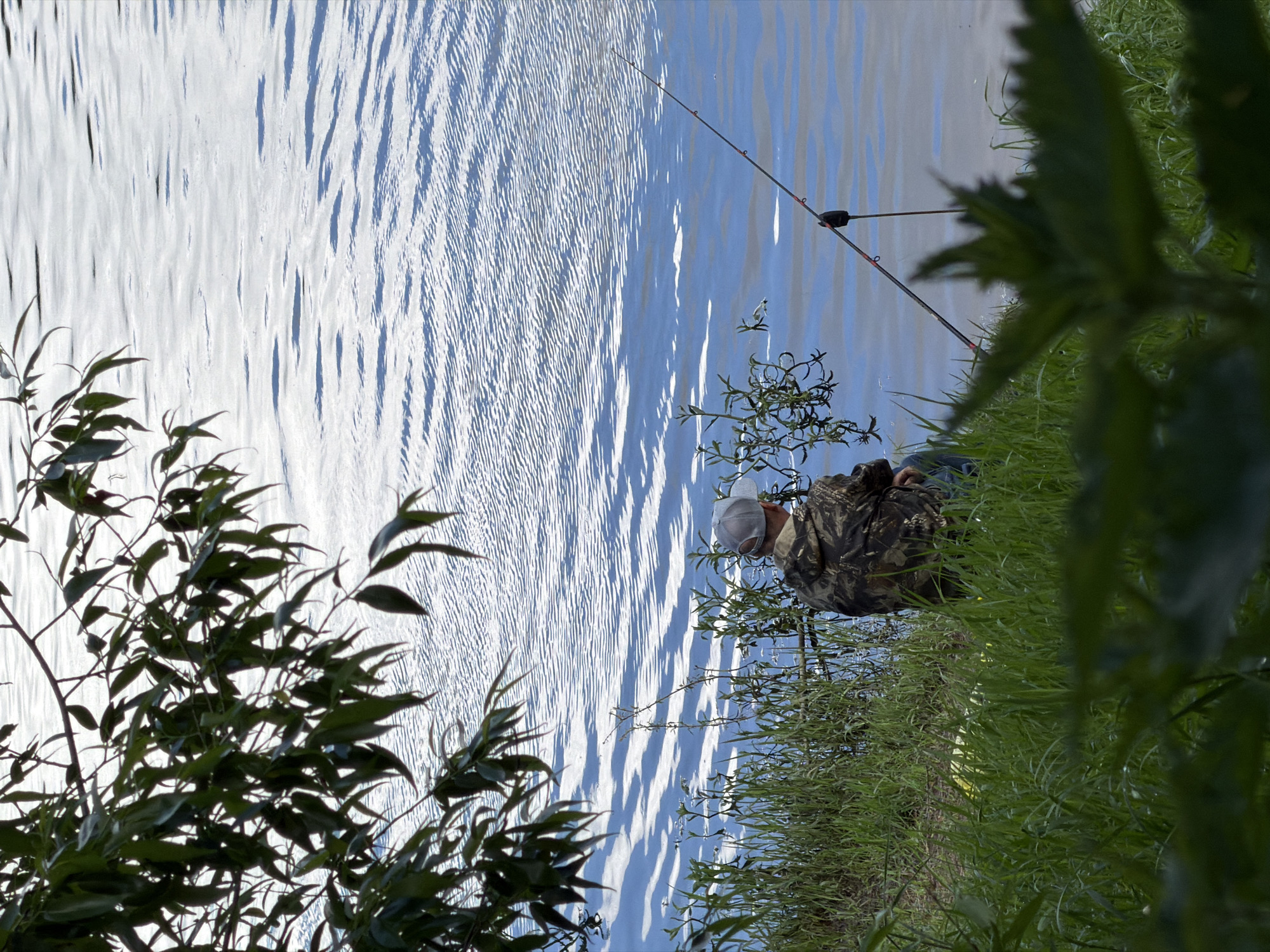 A medium shot shows a person in camouflage clothing fishing by a body of water in an unknown location. The person is wearing a white cap and is positioned to the right of the frame, partially obscured by vegetation. A fishing rod extends out over the water, which reflects a bright sky with a rippled surface. The scene is framed by green foliage in the foreground and along the bank of the water.