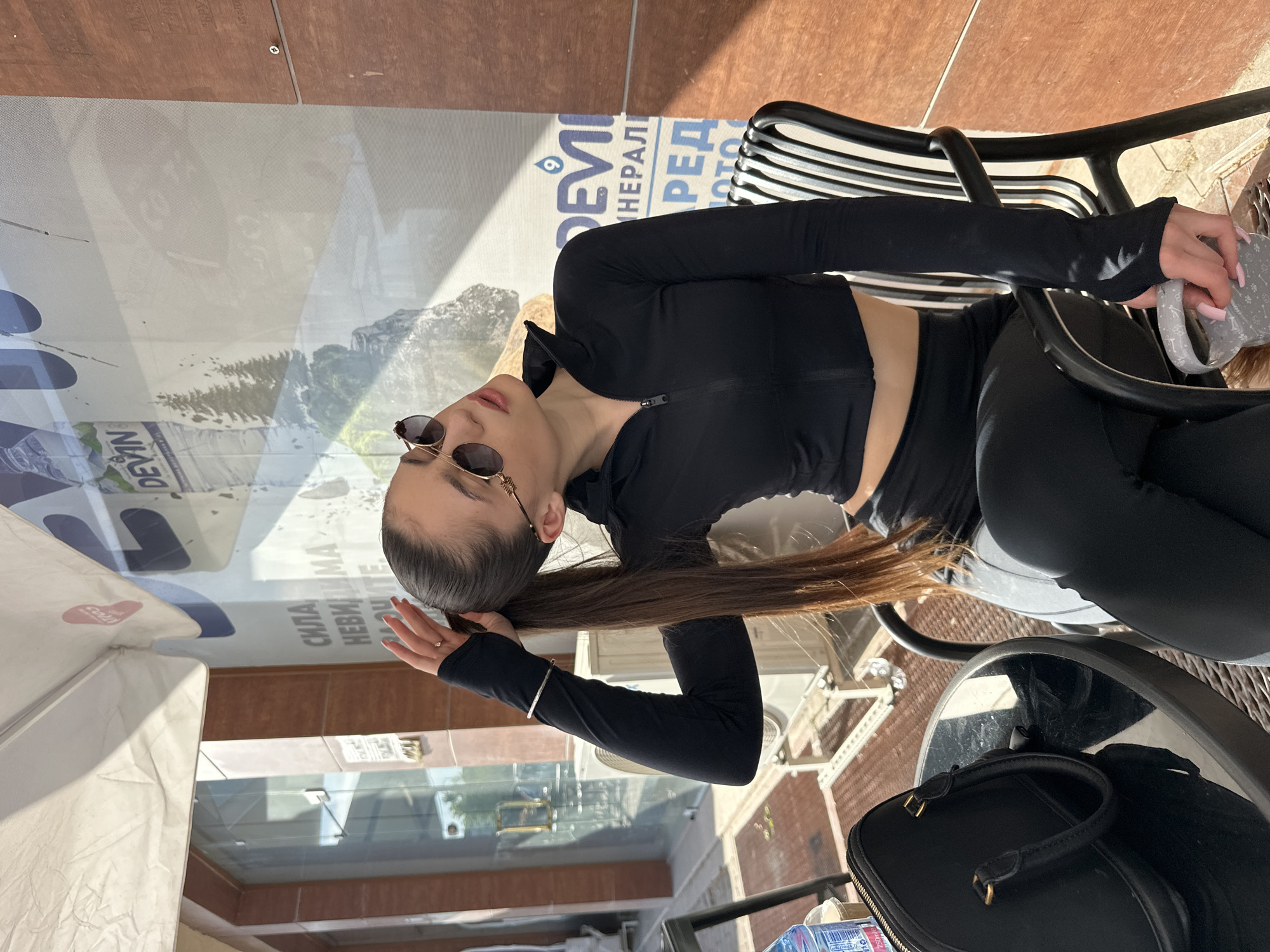 The image, rotated 90 degrees clockwise, shows a young woman seated at an outdoor table, likely at a cafe or restaurant. She has long, dark brown hair pulled back, and is wearing a black, cropped athletic top with a zipper and matching black leggings. Round sunglasses are on her face, and a silver bracelet adorns her left wrist. She holds a grey, patterned cup in her right hand, while her left hand is near her head, which is tilted backward in a relaxed posture.
The setting includes a dark, reflective table and a black slatted chair. On the table next to her, a black handbag or pouch with a gold zipper and handle is visible, along with a partially seen water bottle with a blue label. Behind her, a large glass window or panel reflects a scene featuring mountains, trees, and parts of a building facade.
Prominent Cyrillic text is visible on the reflected signage or banner, aligning with the Plovdiv, Bulgaria location context. Words such as "DEVIN" (a Bulgarian mineral water brand), "МИНЕРАЛНА" (Mineral), "РЕДИ" (Ready/Order), "СИЛА" (Strength), and "НЕВИДИМА" (Invisible) can be discerned. The bright sunlight and distinct shadows suggest it is daytime. The overall scene conveys a casual and relaxed atmosphere.