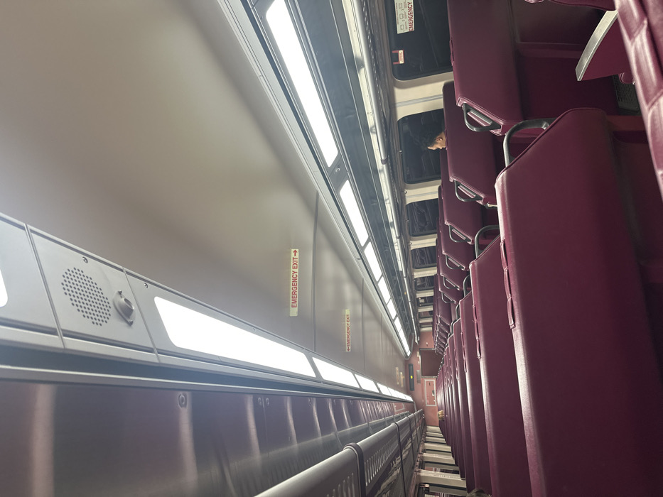 The image depicts the interior of a passenger train car. While the prompt specifies the location as Weston, United States, no visual cues within the image confirm this geographic detail. The perspective is down a central aisle, extending from a low vantage point towards the rear of the car. The ceiling consists of light gray panels, featuring rows of rectangular LED light fixtures providing bright illumination. A circular speaker grille and a small cylindrical sensor are visible on the ceiling toward the upper left. Multiple "EMERGENCY EXIT →" signs are affixed to the ceiling panels, indicating egress routes.
The train car contains numerous rows of seating upholstered in a deep magenta or plum color. These seats are arranged in opposing pairs along both sides of the aisle. One individual is visible on the right side of the car, several rows back from the foreground. This person is seated, facing towards the left (away from the window), with only their head visible in profile. The windows along the sides of the car reveal a dark exterior, indicating either nighttime or transit through a tunnel. Metallic luggage racks are situated above the windows. The overall scene portrays a sparsely occupied, well-lit, and orderly train compartment.