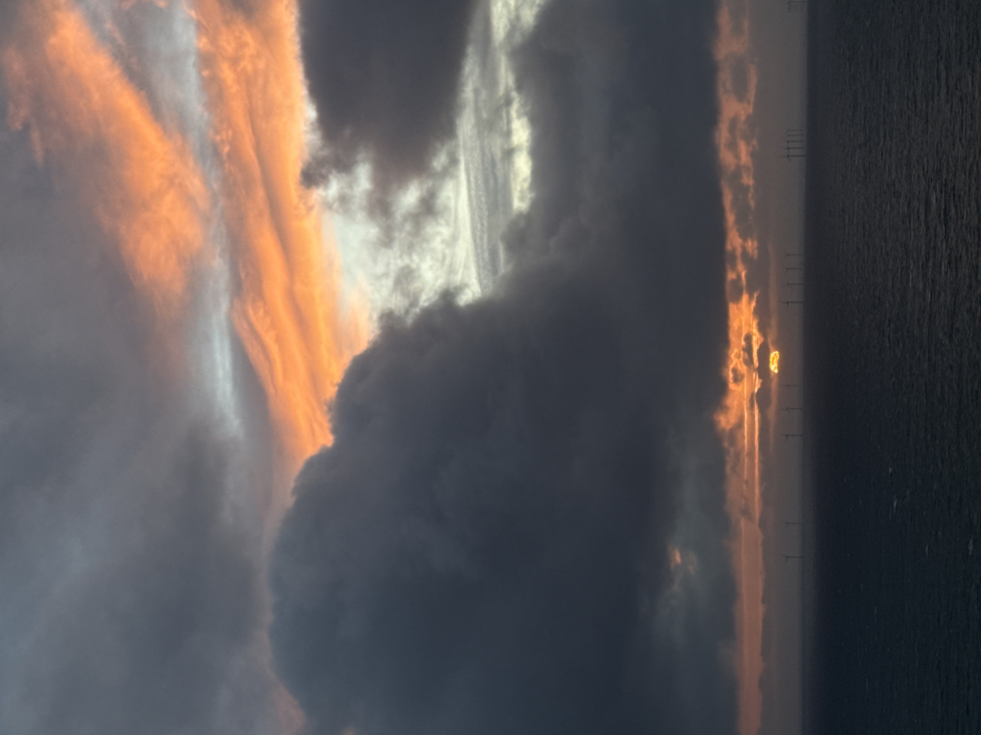 The image captures a dramatic sunset or sunrise over the sea, featuring an offshore wind farm on the horizon. The sky, which occupies the majority of the frame, is filled with an impressive array of clouds. On the left side, the clouds are intensely illuminated by the low-lying sun, showcasing vivid streaks of orange, yellow, and reddish hues that blend into soft whites and light grays, suggesting complex atmospheric layering. Towards the center and right, the clouds become denser and darker, appearing in deep grays and bluish-blacks, indicating thick storm clouds or areas where the light is entirely obscured.
The horizon line spans across the lower portion of the image, separating the dynamic sky from the water. The sea, visible on the right, is a dark, muted blue-grey, with a gently textured surface. Along the horizon, numerous tall structures, identifiable as wind turbines from an offshore farm, stand silhouetted against a narrow band of golden light. The sun's intense glow, possibly a partial view or a bright reflection, can be seen just above the horizon, peeking through a gap in the clouds and casting a brilliant yellow-orange light that highlights the cloud edges and reflects faintly on the water.
The scene conveys a powerful and awe-inspiring natural moment, characterized by the dramatic interplay of light and shadow across the vast sky and sea. The presence of an offshore wind farm aligns with the provided location context of Fécamp, France. No people or visible text are present in the image.