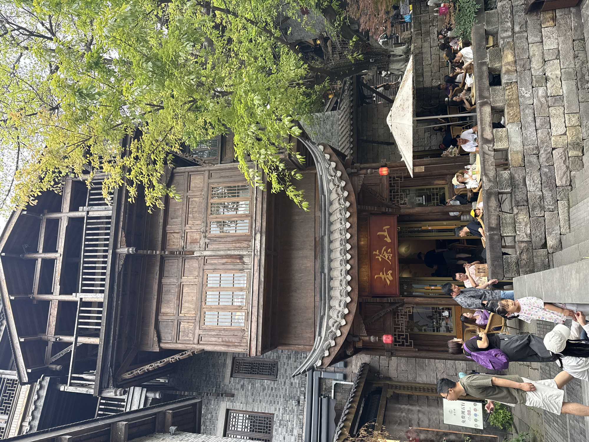 The image depicts a bustling scene in Yuzhong District, China, in an area with traditional architecture. A multi-story wooden building with intricate woodwork details, including a projecting bay window and a curved roof, dominates the left side of the image. The structure is likely a restaurant, as suggested by the sign with Chinese characters above its entrance. People are seated at tables outside the restaurant, some dining, while others are walking along the adjacent stone pathway. A large tree provides shade over the scene. The street is constructed of grey stone blocks. Overall, the image captures the vibrancy of a popular area within Yuzhong District.