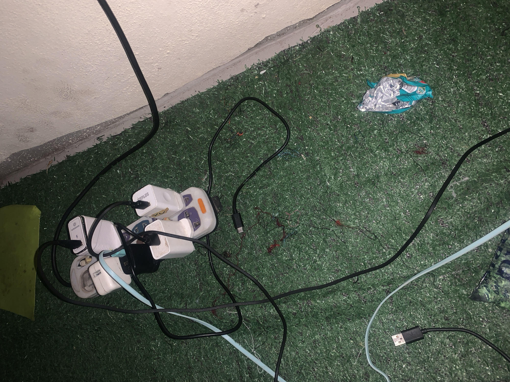 This is a medium-angle, eye-level shot of an electrical surge protector and multiple charging adapters, all plugged into each other and connected by various black and light blue cables. The items are spread across a green, artificial turf-like surface, which appears to be the floor of an outdoor or semi-outdoor space, possibly a patio or balcony. In the background, a textured, light-colored wall runs diagonally across the frame. To the right of the surge protector, a crumpled silver and teal object, possibly a snack wrapper, lies on the artificial turf. The overall impression is one of disorganization and a potential fire hazard due to the tangled wires and multiple devices. The location is identified as Jalingo, Nigeria.