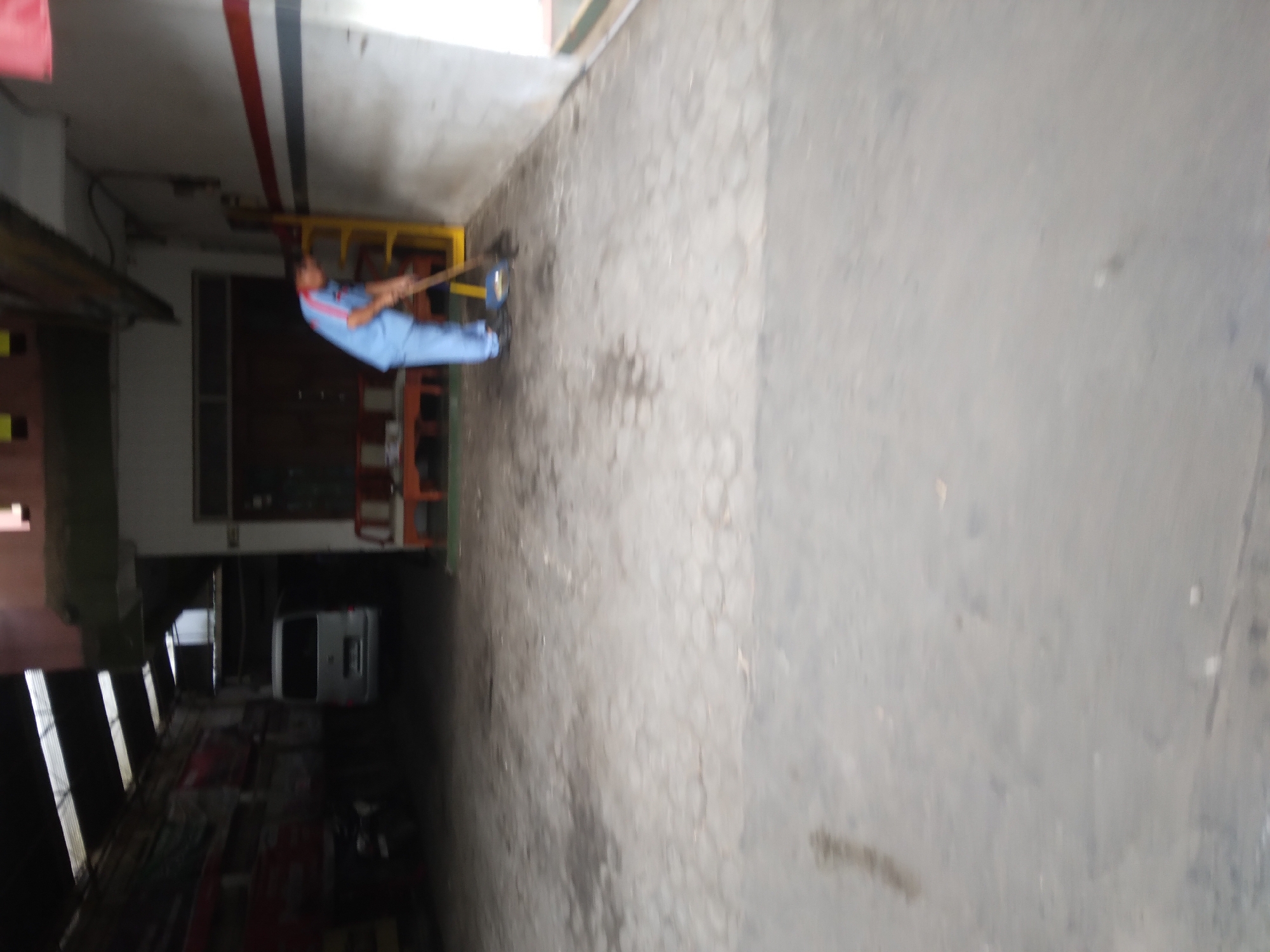A person wearing a light blue uniform is sweeping the ground with a broom. The person is standing on a grey paved surface, which appears to be a pathway or a driveway. Behind the person, there is a white wall with red and dark grey stripes. To the left, there is a dark interior space with a roof supported by beams, and a white van is visible parked inside. Further to the left, a motorcycle is parked. In the background, a yellow ladder leans against the wall. The scene appears to be an outdoor or semi-outdoor area within a facility, possibly in Berkoh, Indonesia, given the context.