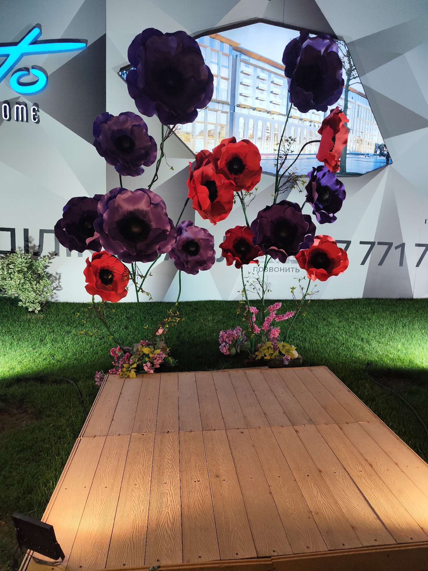 The image shows a retail or event display featuring large, artificial poppy flowers in vibrant red and deep purple. The flowers are arranged on thin, dark stems, creating a dramatic visual effect against a grey and white geometric backdrop. Behind the flowers, a large screen displays an architectural rendering of a modern building with glass facades and arched entrances. The screen is framed by the geometric pattern of the wall.
The foreground consists of a wooden platform, appearing to be a stage or display area, illuminated by warm spotlights from the left. The platform is constructed from light-colored wooden planks with visible wood grain and nail heads. The platform is set on a vibrant green lawn, which is also illuminated, suggesting an outdoor or indoor setting designed to mimic an outdoor space. Small clusters of artificial flowers in pink and yellow hues are placed at the base of some of the poppy stems, near the platform.
On the left side of the backdrop, a blue neon sign with the stylized letters "to" is visible, and below it, the word "home" in white block letters. Further to the right on the backdrop, a phone number appears in large grey numerals: "7717". A portion of text, likely in Cyrillic, that reads "позвонить" (which translates to "to call") is visible in smaller grey lettering above the wooden platform and below some of the flowers.
The lighting is a mix of cool, bright light illuminating the lawn and warm spotlights focused on the wooden platform, creating a contrast in the scene. The overall impression is one of a modern and decorative display, possibly for a real estate or retail promotion, with a clear emphasis on visual appeal.
The provided information states the location is in Almaty, Kazakhstan. However, there are no specific landmarks or visual cues in the image that definitively place it within Almaty. The architectural rendering on the screen is generic and doesn't point to a specific building in the city. The text on the backdrop, "7717" and "позвонить," are common elements for contact information and do not offer specific geographical clues. The neon sign "to home" is also a generic branding element. Therefore, while the context provided is Almaty, Kazakhstan, the image itself does not contain irrefutable proof of this location.