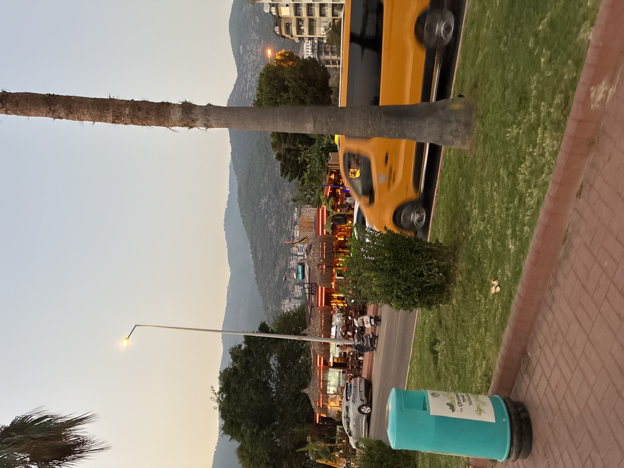 A street scene in Alanya, Turkey, captures a coastal town atmosphere at dusk. The foreground features a turquoise trash receptacle on a brick sidewalk, adjacent to a grassy area. A palm tree trunk stands prominently in the middle right of the frame. Across the street, a row of restaurants with thatched roofs are illuminated, suggesting evening dining. Several scooters are parked along the sidewalk in front of the establishments. A silver car is parked on the street, and a motion-blurred yellow van, possibly a taxi, drives by on the road. In the background, lush green hills rise, with buildings dotted along their slopes, indicating the city's urban sprawl. The sky is a soft gradient of pale yellow and blue, typical of sunset or early morning light.