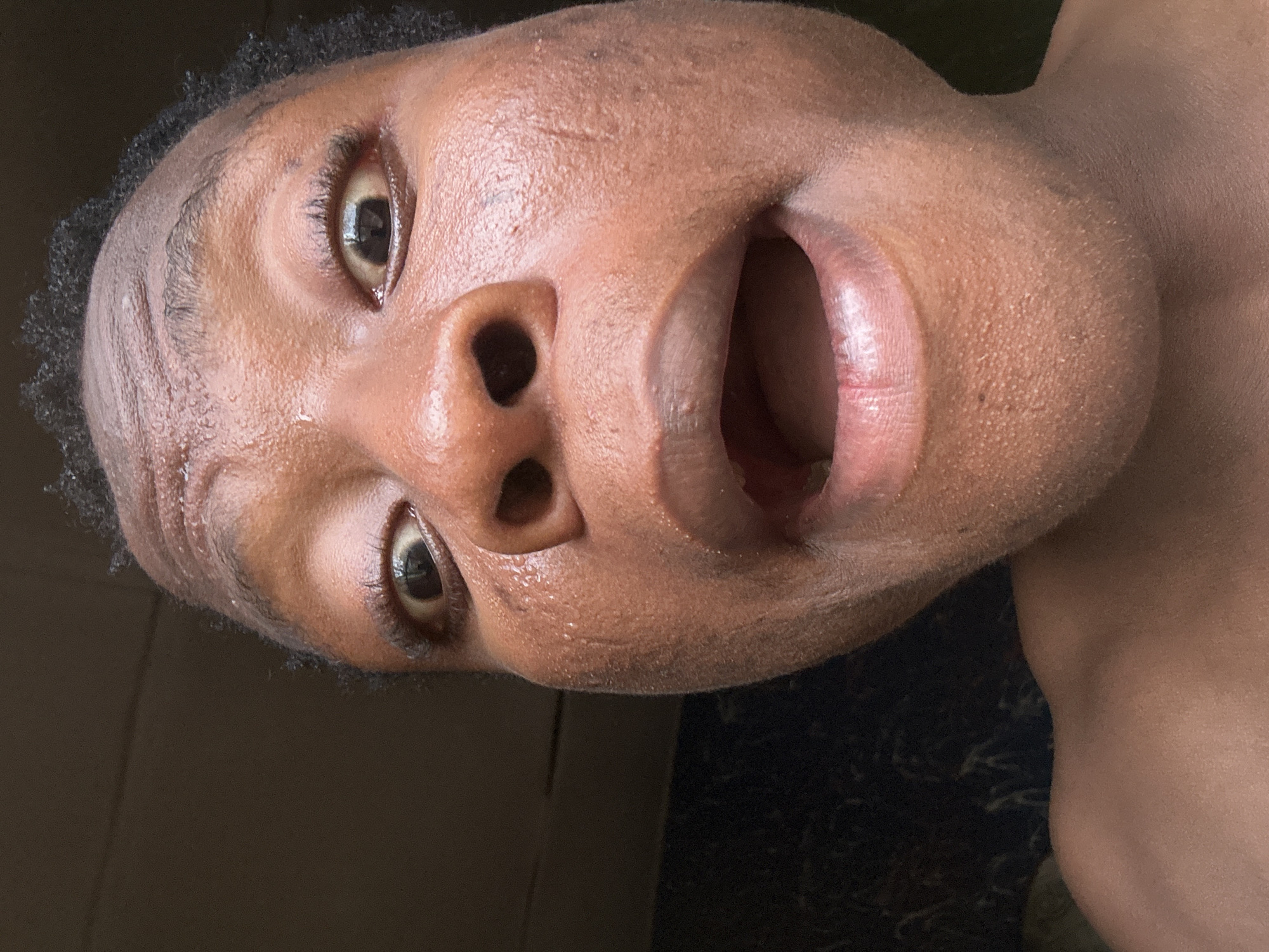 Close-up of a Black person's face with an open mouth, appearing to be yelling or singing. The skin is slightly shiny, possibly due to sweat. The background is dark and indistinct. The location is unknown.