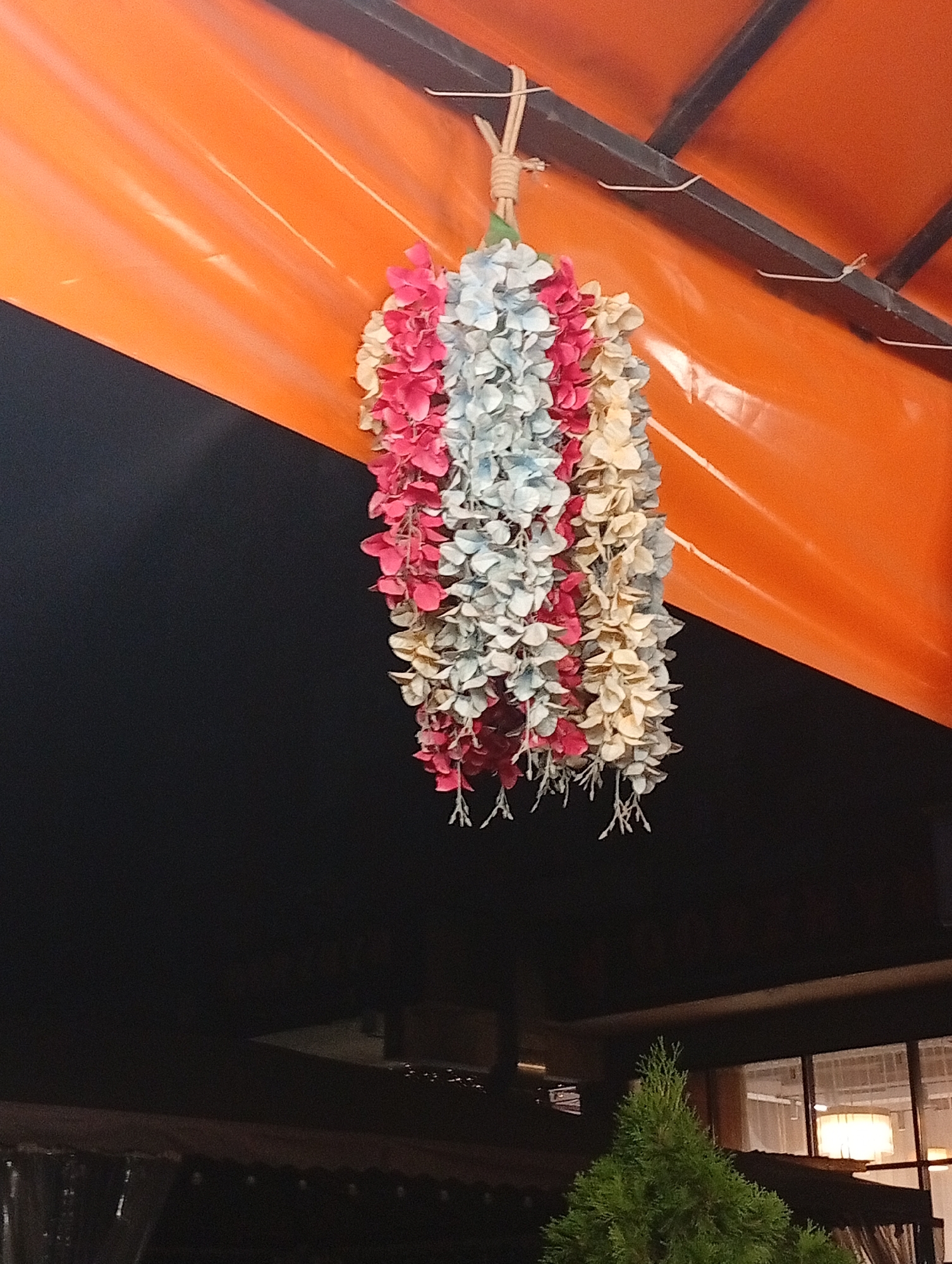 A hanging decoration made of faux flowers in shades of pink, blue, and cream is suspended from a metal frame against an orange awning. The awning is supported by dark metal beams, and below it, a dark, out-of-focus background is visible. In the lower portion of the image, a dark roof structure is present, with a green evergreen tree and a building with windows illuminated by interior lights visible beyond it. The scene appears to be an outdoor setting, possibly a cafe or restaurant, though no specific location in Almaty, Kazakhstan, can be determined from the visual information provided.