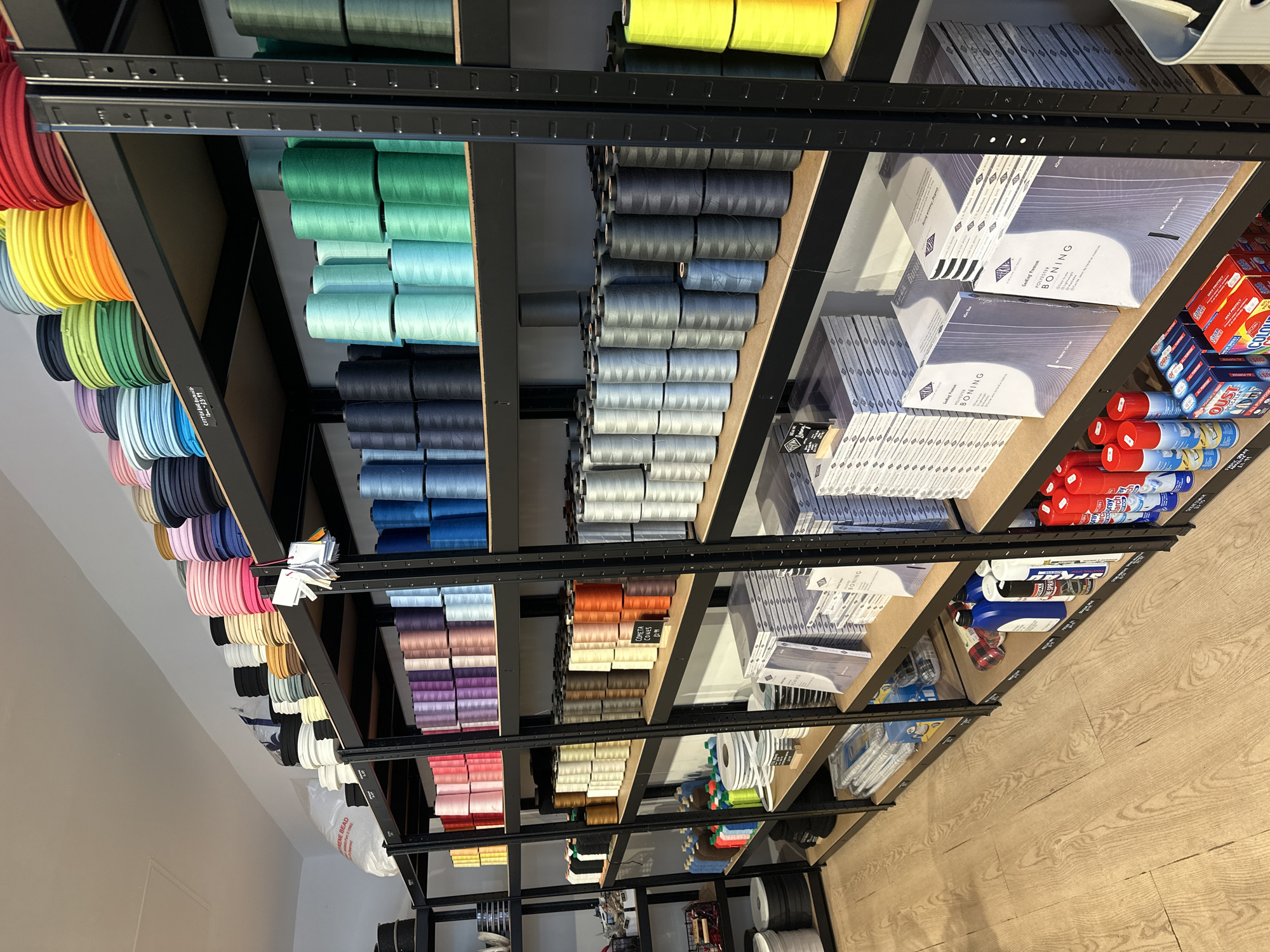 The image shows a retail display shelf, likely in a craft or sewing supply store. The shelves are black metal, and contain various items neatly arranged. The first two shelves are filled with spools of thread or ribbon in a wide range of colors, including reds, oranges, yellows, greens, blues, and purples. There are stacks of neatly arranged boxes and cartons on the remaining shelves, indicating a diverse product selection. Various cleaning products are stored next to the cartons. The store appears well-organized. The OCR indicates that the image includes a label reading 'RENE BEAD,' and indicates the products are Polyester Boning, Cotton Bus Binding and Cometa Cones, as well as various spray cleaning products. The location is likely the city of London, United Kingdom.