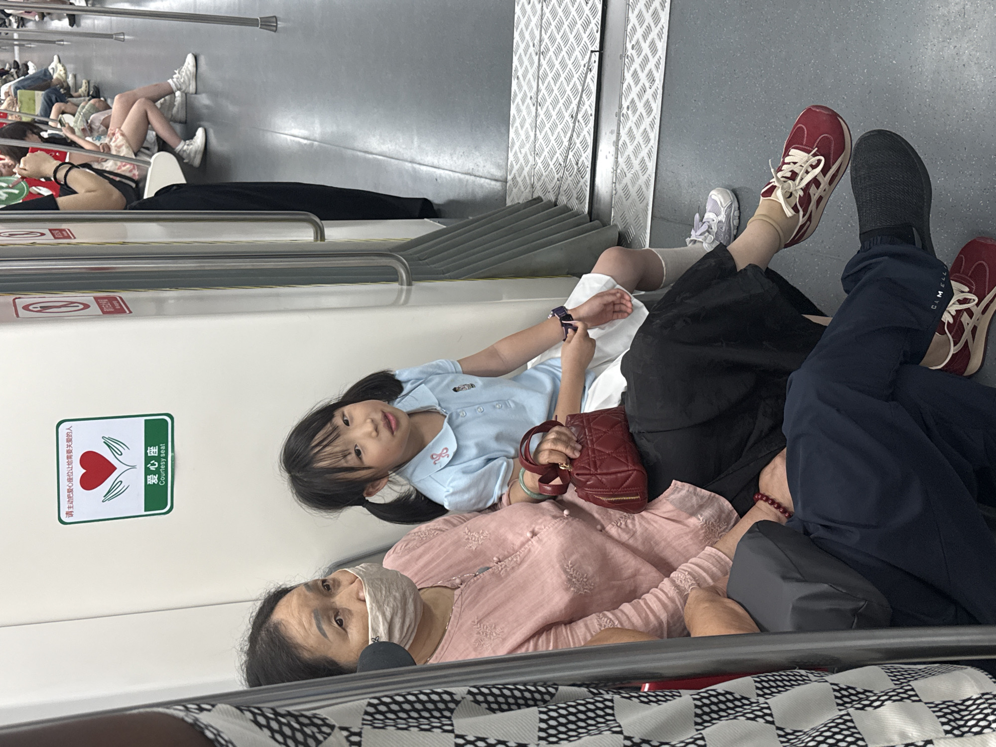 The scene depicts a group of individuals resting on the seats of a train in Yuzhong District, China. An elderly woman wearing a light pink shirt and a face mask is lying down. Next to her is a young girl, resting on her side, wearing a light blue polo shirt, and holding a small, maroon-colored purse in her left hand. An adult wearing a pair of dark grey shoes and blue pants with a burgundy shoe, is seated and has their legs stretched to their side. Other individuals, sitting with their legs out, are visible further down the train car. A courtesy seat sign is posted on the wall.