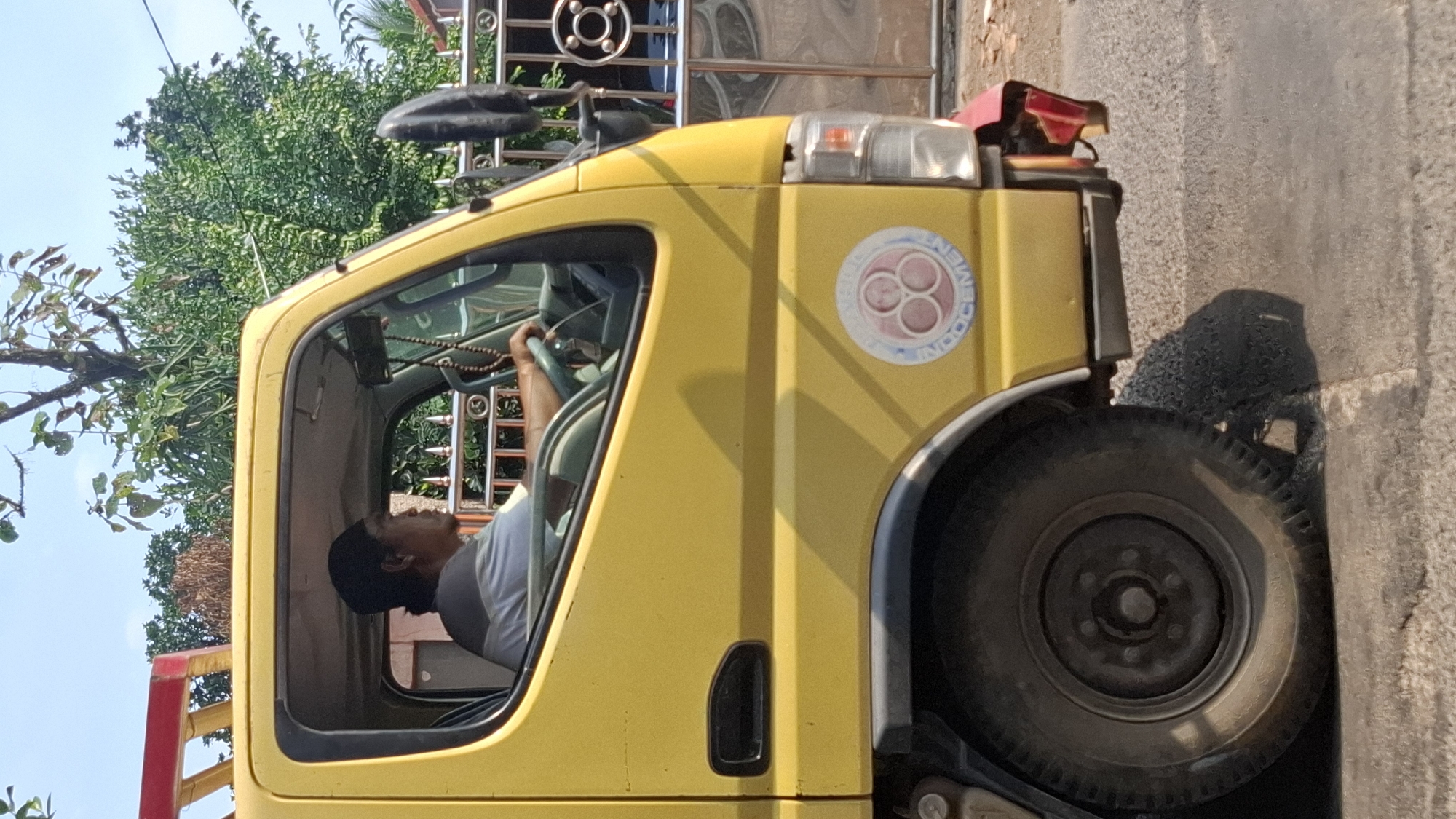 A side profile of a yellow truck is visible, with the driver, a man wearing a dark cap and a light-colored shirt, seated in the cabin. He is holding the steering wheel, and his right arm is resting on it. The truck has a black side mirror and a circular emblem on its side door featuring interconnected rings and the word "INDOCEMEN". The front wheel of the truck is partially visible, and a shadow is cast on the road beneath it. The background shows lush green trees and a blue sky, with a portion of a metal fence and a building also discernible. The setting appears to be a roadside in Cilaku, Indonesia, indicated by the visual cues of vegetation and architecture common to the region.