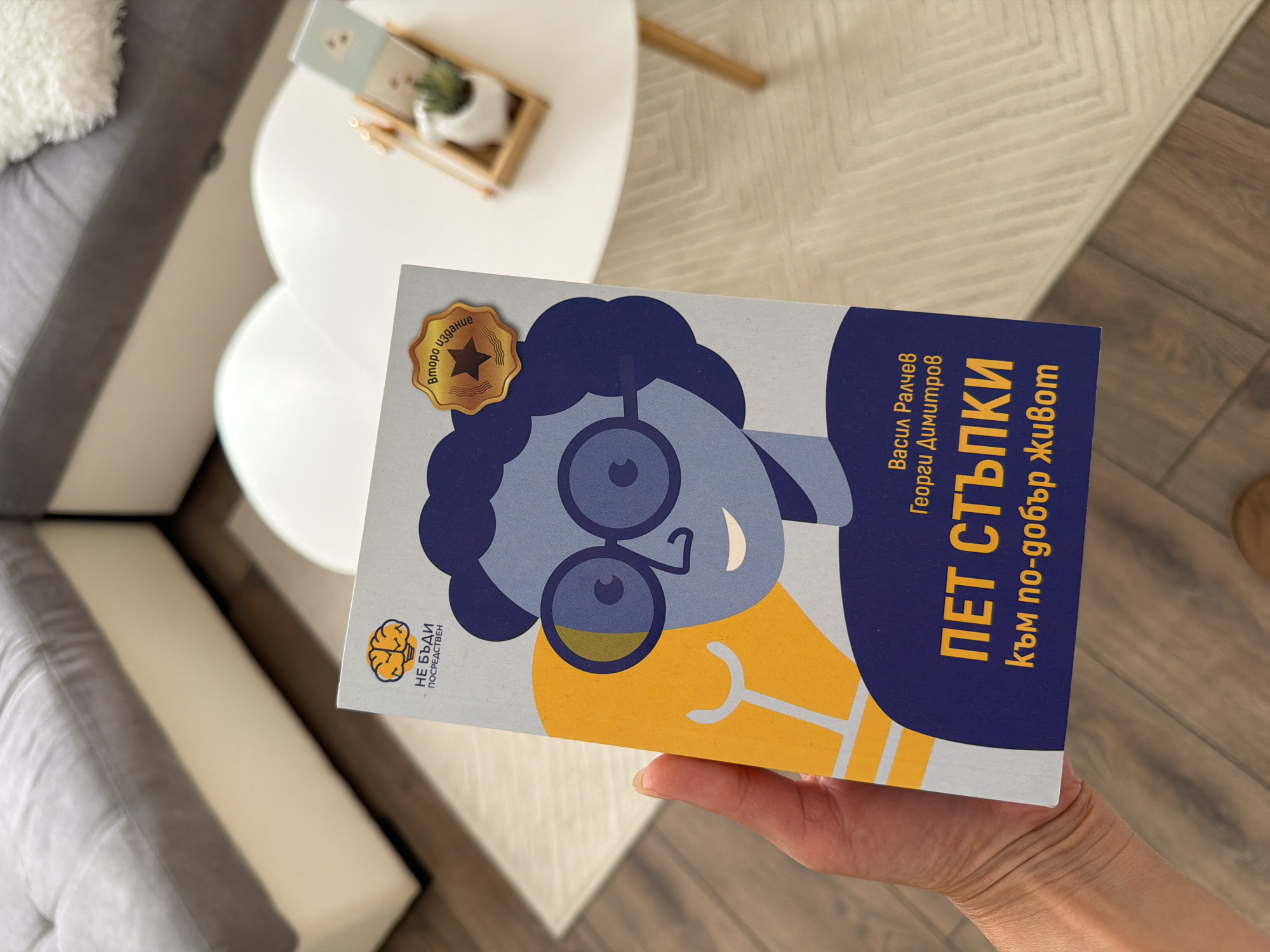 An overhead shot shows a person's hand holding a book with a blue and yellow cover, angled towards a white, cloud-shaped coffee table. The book's cover features a stylized blue illustration of a person wearing glasses and a yellow lightbulb graphic. Text on the cover is in Bulgarian and reads "ПЕТ СТЪПКИ към по-добър живот" (Five Steps to a Better Life), with author names "Васил Ралчев" and "Георги Димитров" above it. A small, gold, star-shaped emblem is on the upper right corner of the book, with the text "Второ издание" (Second Edition). The coffee table has a small wooden tray with a white pot and a succulent plant. In the background, a section of a grey and white sectional sofa is visible. The scene appears to be indoors, likely in a living room, with a light-colored wooden floor. No elements in the image suggest a specific location within the city of Pazardzhik, Bulgaria.