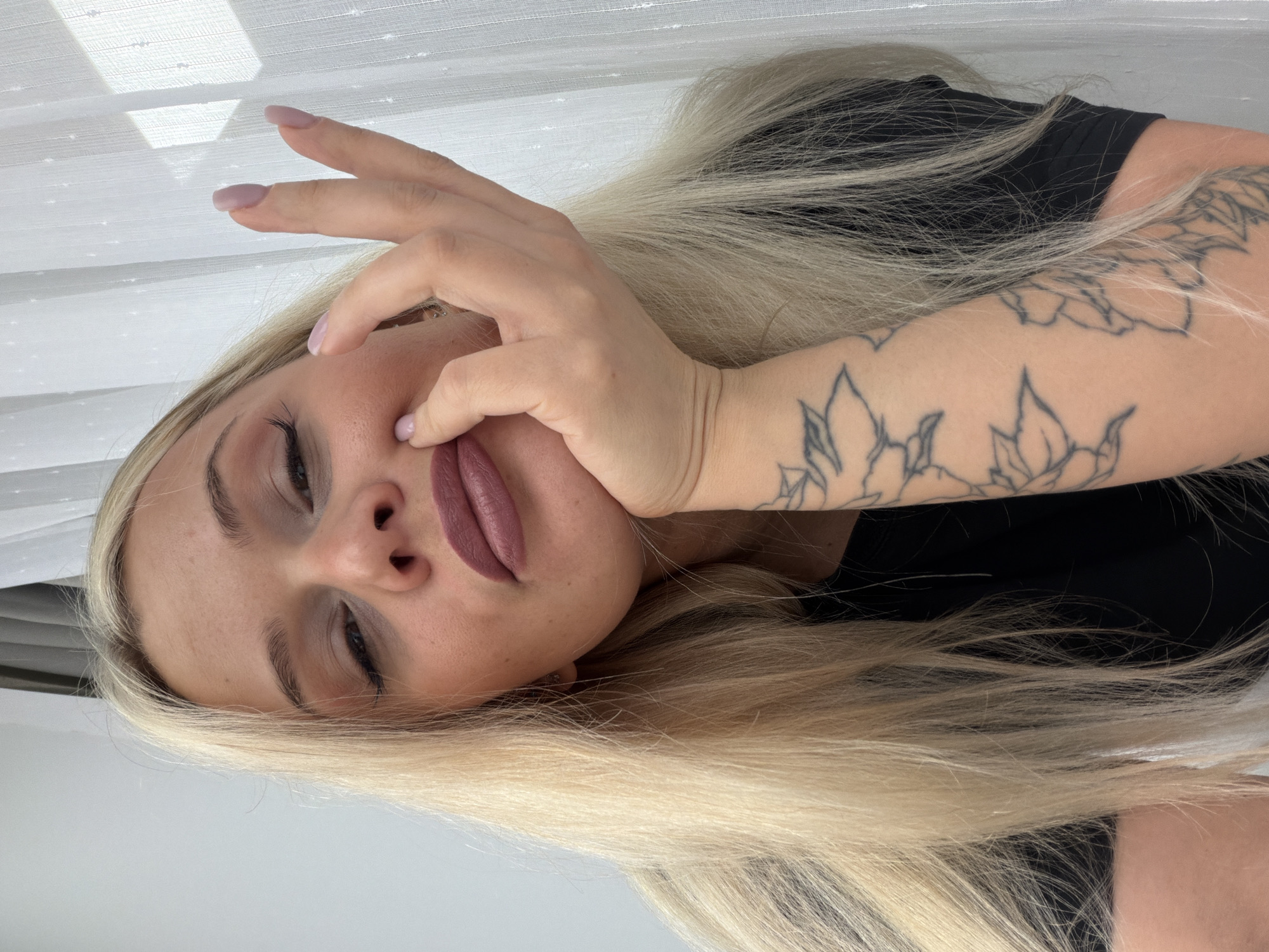 A portrait-style image features a young woman with long, straight blonde hair, positioned with her head tilted to the left. She has light skin, prominent eyebrows, and is wearing eye makeup and a matte, mauve-pink lipstick. Her left hand is gently placed against her face, with her fingers lightly touching her cheek, nose, and lips. Her fingernails are manicured with a soft pink polish. Floral or leaf outline tattoos are visible on her left forearm. She is wearing a dark top, likely black. The background on the left side shows a white, possibly sheer, curtain or window blind with horizontal lines and shadows, suggesting natural daylight. The rest of the background is a plain, light surface. The woman's expression is calm and neutral, possibly contemplative. No text is visible in the image. The provided location context for this image is Sarapul, Russia.