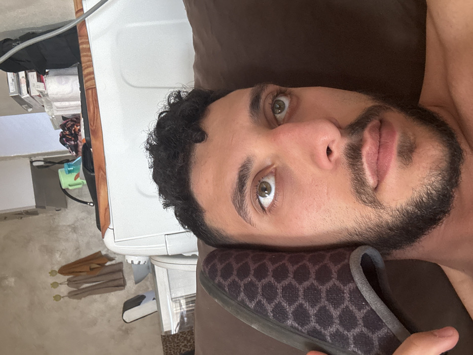 A male individual with dark curly hair, a beard, and light-colored eyes is depicted holding a dark, honeycomb-patterned slipper to his left ear, gazing upwards. He is shirtless and appears to be reclining on a brown leather or faux-leather surface, likely a couch or cushion. The background reveals an indoor setting with a textured, light-colored wall. Three beige towels hang on yellow hooks to the left. Below them, a small white appliance or container is next to a light gray bowl. A large white appliance, possibly a washing machine, is visible in the mid-ground, topped with a green cup, a blue bottle, patterned fabric, rolls of white paper, stacks of white boxes, and a black bag. A gray cable is visible in the upper right quadrant. The scene is located in the city of Gradishte, Bulgaria.