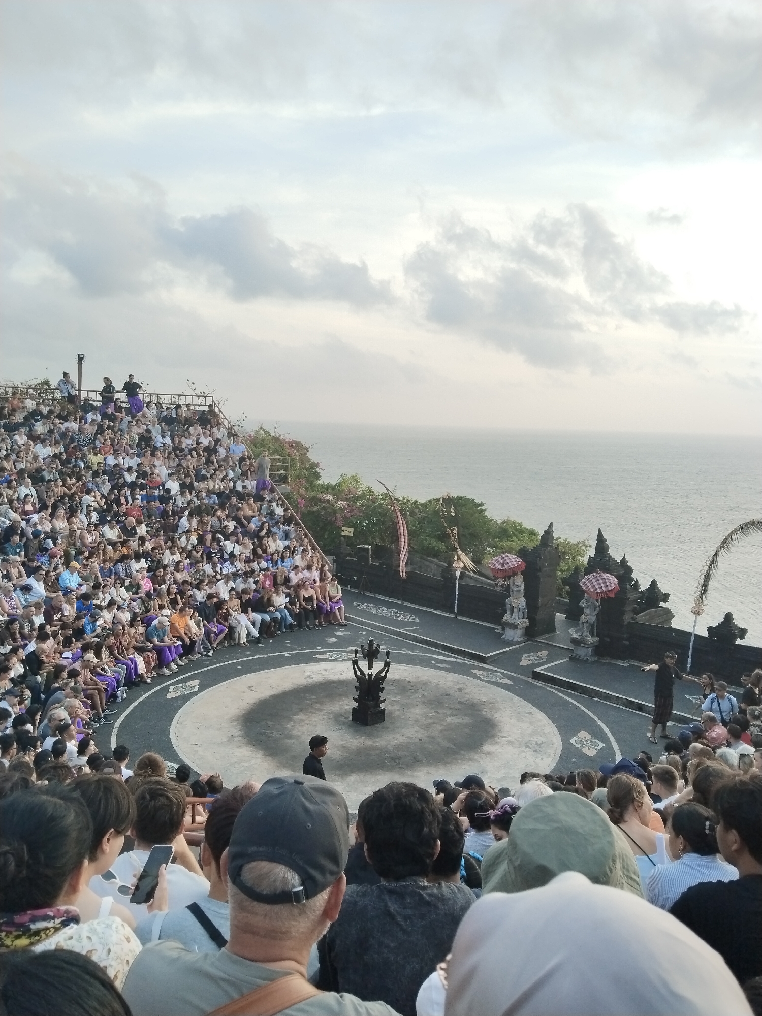 An aerial view captures a large outdoor gathering, possibly a performance, in Kuta Selatan, Indonesia. A vast audience is seated on tiered stone steps overlooking a circular performance area. The ocean forms a backdrop to the scene, with a hazy sky above. The performance area features a dark, ornate sculpture at its center. Several individuals are visible within the performance space, including one person in a black shirt and sarong. Statues and decorative elements, including red and white checkered umbrellas, are placed along the edge of the performance area. The audience, a diverse mix of individuals, fills the seating tiers, many appearing engaged with the performance. Some people in the foreground are looking towards the performance area, with one person holding a mobile phone, likely capturing the event. The overall atmosphere suggests a cultural or tourist event.