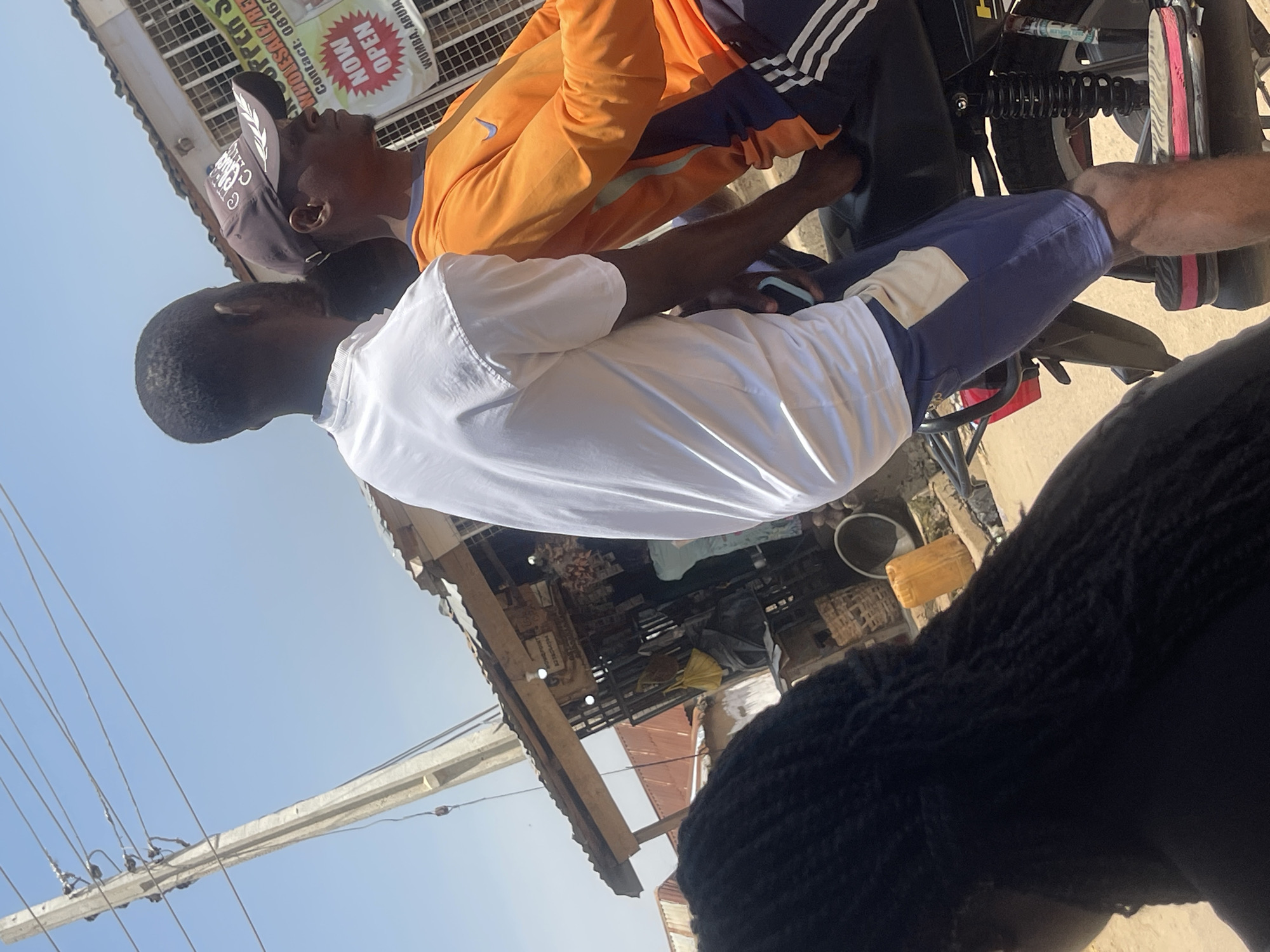 The image shows two men riding a motorcycle in Abuja, Nigeria. The man in front is wearing an orange long-sleeved shirt and a dark baseball cap. The man behind him is wearing a white t-shirt and blue shorts. A woman with long black hair is partially visible in the foreground, to the left of the motorcycle. Behind the motorcycle, there is a small market stall with a corrugated metal roof. A sign on the stall reads "NOW OPEN" and displays a contact number. The scene is outdoors, with a clear blue sky and a power line visible in the background. The ground appears to be dirt or gravel.