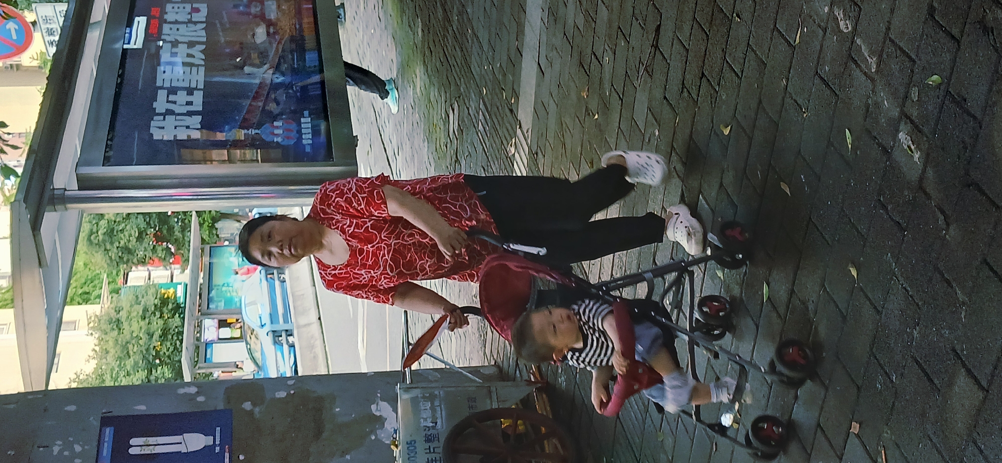 A daytime scene shows a woman in a red patterned shirt and black pants walking along a sidewalk in Shapingba District, China. She is pushing a red stroller containing a child wearing a striped shirt and light-colored pants. The sidewalk is paved with gray brick. Beside them is a bus shelter featuring an advertisement with large white Chinese characters that translate to "I am in Chongqing." In the background, there is a street with vehicles and greenery.