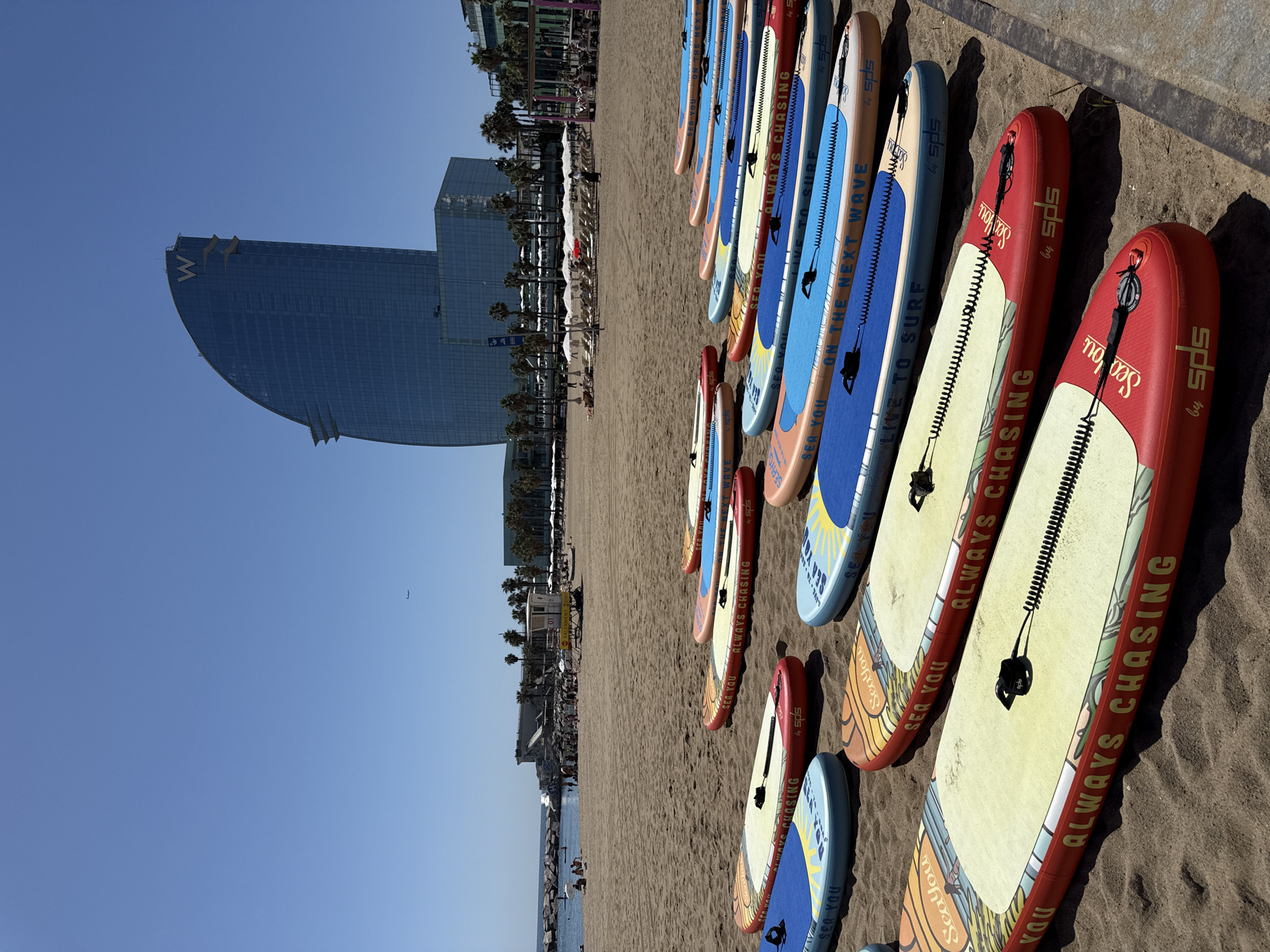The image captures a sunny day at the beach in Barcelona, Spain, likely on Barceloneta Beach. In the foreground, numerous inflatable stand-up paddleboards are arranged on the sand, some with colorful graphics and text such as "ALWAYS CHASING" and "SEA YOU." In the background, the iconic W Hotel, a sail-shaped skyscraper, dominates the skyline. Beyond the paddleboards, people are visible on the beach and in the shallow water, with umbrellas and loungers set up. The sea stretches to the horizon under a clear blue sky, with a small airplane visible in the far distance. The sand is light brown and appears to be somewhat wet near the water's edge. Palm trees and other buildings line the beachfront, suggesting a lively urban coastal environment.