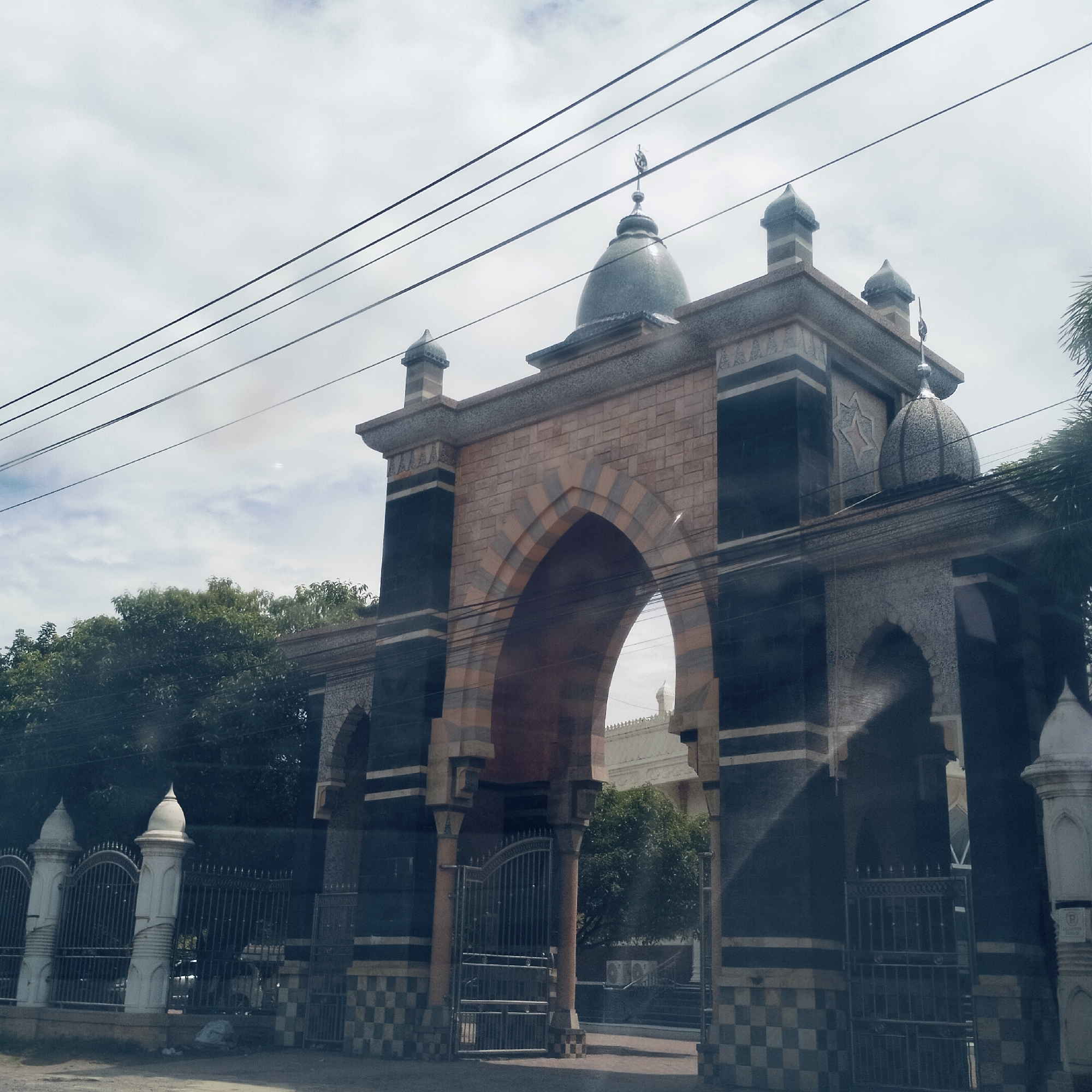 The image features a grand, ornate gate structure, likely serving as an entrance to a mosque or Islamic complex in Cotgadong, Indonesia. The gate exhibits prominent Islamic architectural elements, including several pointed arches. The largest central archway is constructed from alternating light and darker reddish-brown stone blocks, creating a striped pattern.
The main pillars of the gate are clad in dark, possibly polished, panels, accented with lighter horizontal bands and geometric star motifs. The base of the structure features a checkered pattern of light and dark tiles. The gate is topped with multiple domes: a large, greenish-blue dome with a finial incorporating a crescent moon and star symbol sits centrally, flanked by smaller, pointed domed turrets. Another smaller, intricately patterned dome is visible on the right side.
Overhead, multiple power lines crisscross the frame against a bright, cloudy sky, partially obscuring the upper details of the structure. Lush green trees are visible behind and to the sides of the gate, suggesting a natural environment within or beyond the complex. To the left, a decorative metal fence with ornate white pillars extends from the gate. Through the main archway, a portion of a lighter-colored building, possibly the mosque itself, can be seen in the background.
The scene depicts a static, daytime view with no people or specific activities visible. The architectural style strongly suggests a cultural or religious landmark.