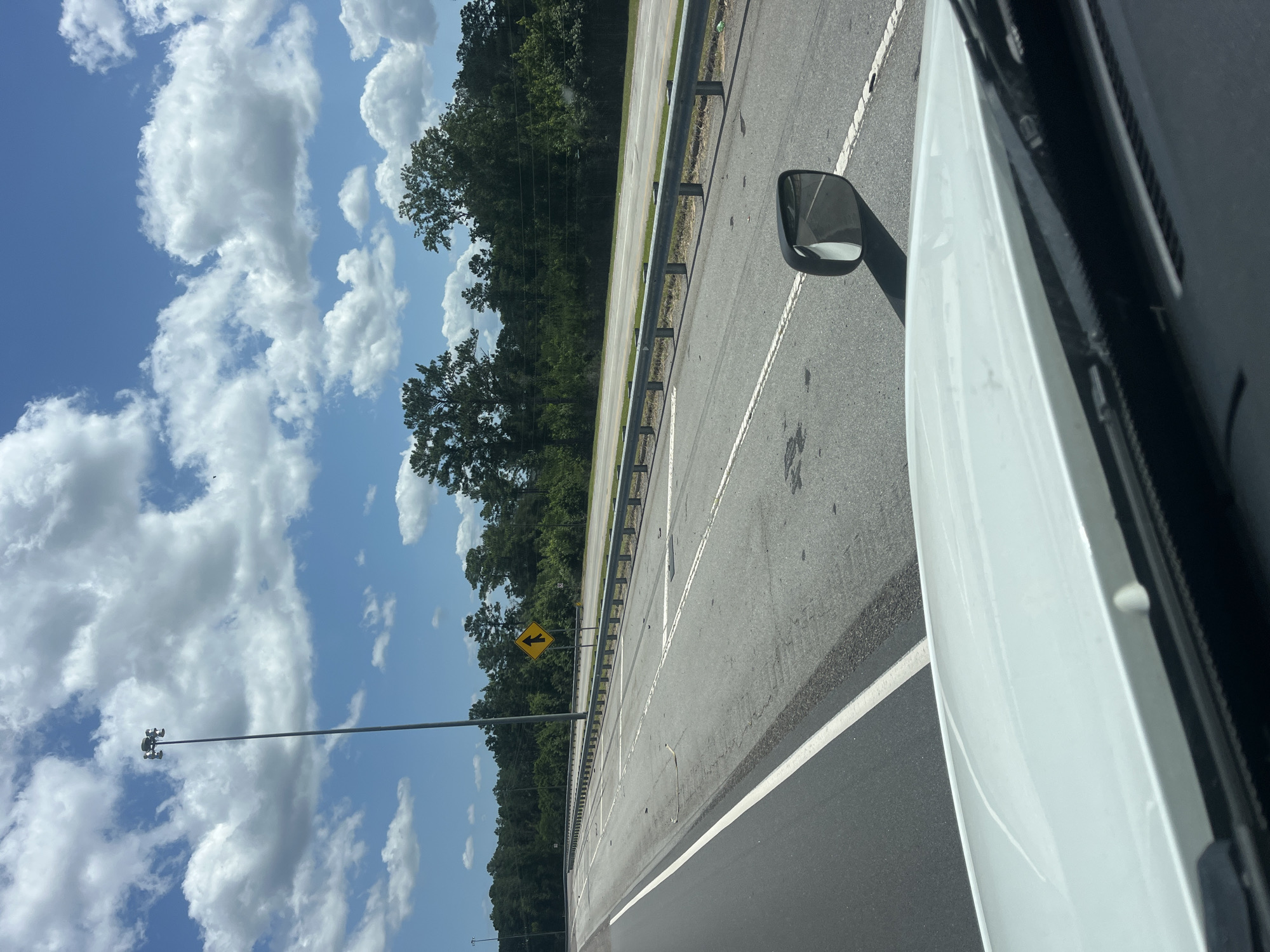 The image depicts a highway scene in Riceboro, United States, under a partly cloudy sky. The perspective is from inside a white vehicle, showing the hood and windshield wipers. The highway itself has multiple lanes, marked with white lines. A metal guardrail runs along the side of the road, separating it from a wooded area in the background. A tall light pole with multiple lamps is visible on the left side of the highway. A yellow diamond-shaped road sign with an arrow pointing upwards is positioned near the guardrail. Scattered debris can be seen on the road surface. A side mirror of the vehicle is also visible, reflecting the highway and the sky.