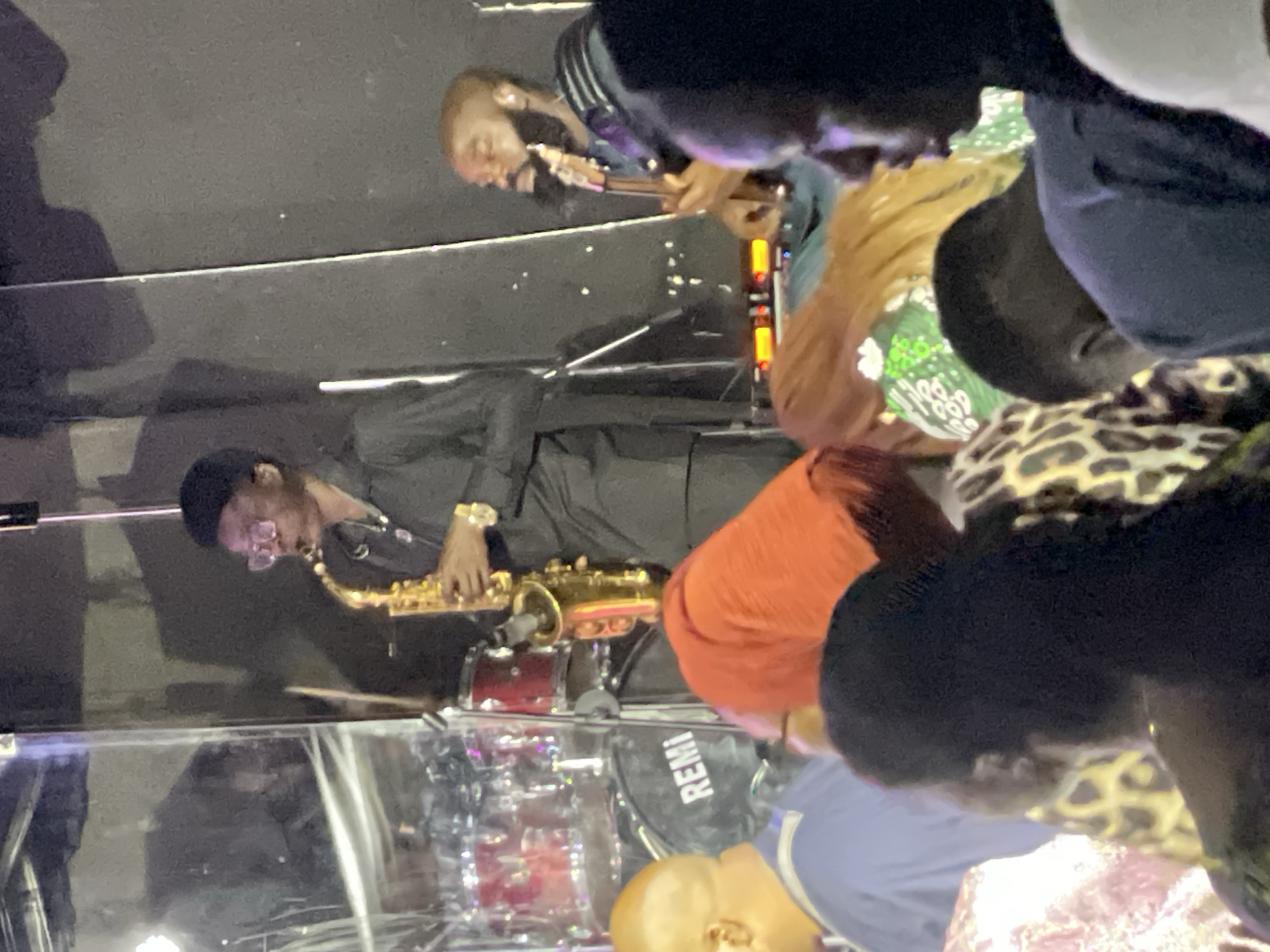 A live music performance is underway, featuring a saxophone player and a guitarist on a stage in an indoor venue. The saxophone player, dressed in a black suit and cap, is actively playing his instrument. The guitarist is positioned behind him. A drum set is visible. A crowd of people is in attendance, obscuring parts of the performance. The location of the image is in the city of Jos, Nigeria.
