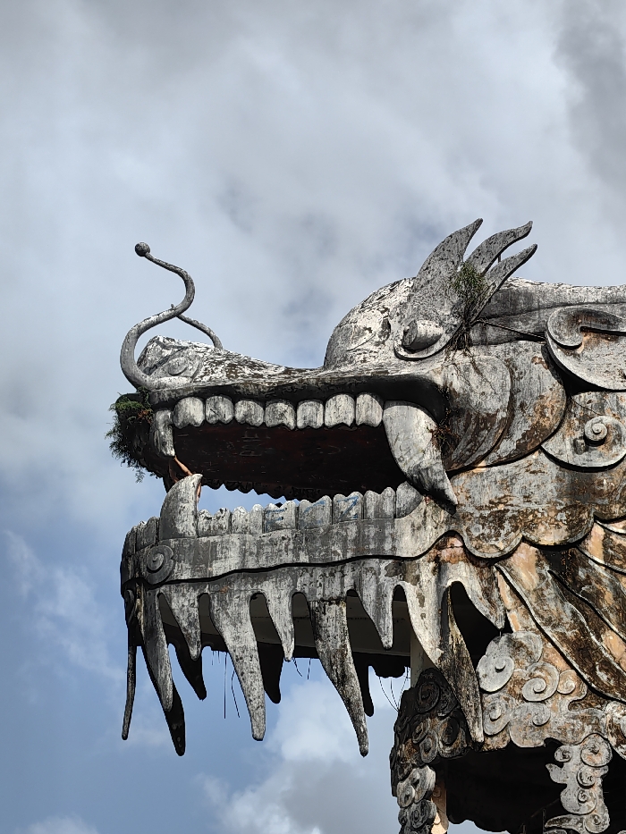 The image displays a weathered concrete sculpture of a dragon's head and upper body, located in Thủy Xuân Ward, Vietnam. The dragon, depicted with its mouth agape, is primarily grey and off-white, showing extensive signs of exposure and deterioration, including staining, minor structural damage, and the growth of small green plants and moss in its crevices. Its head features intricate scale patterns, prominent pointed teeth lining both the upper and lower jaws, and two distinctive curled, antenna-like structures extending from its snout, each terminating in a spherical tip. Three fin-like appendages are visible on the right side of its head. The interior of the mouth appears dark red-brown. The sculpture is set against an overcast sky, with varying shades of grey and white clouds. No people or active elements are present in the scene.