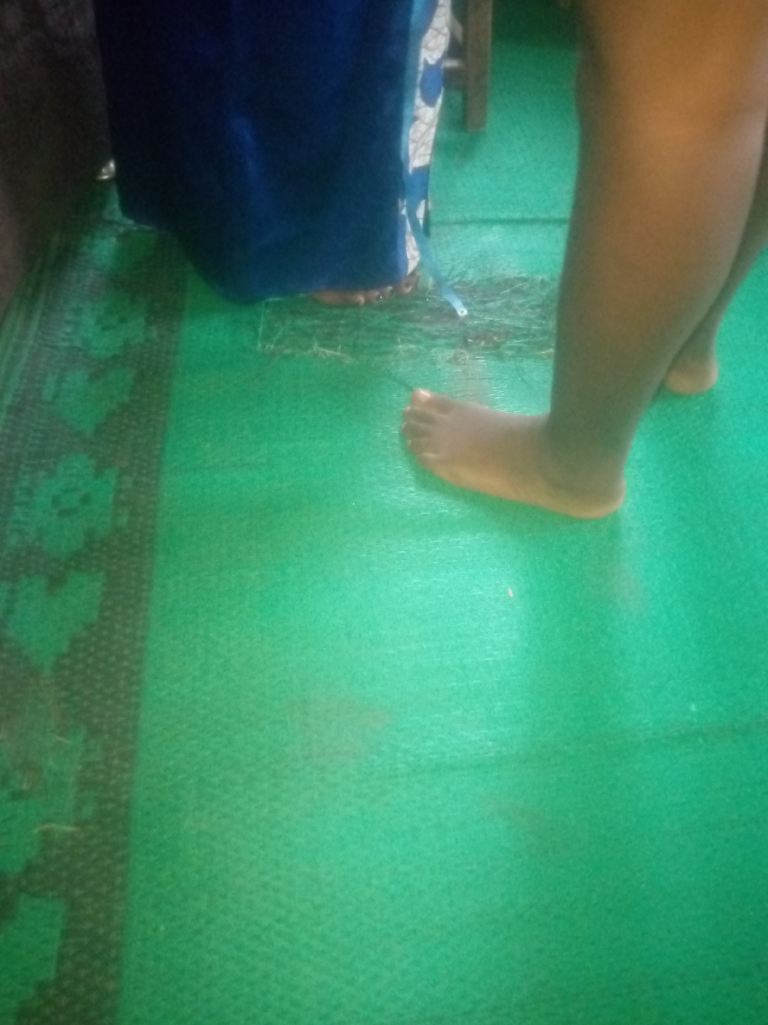 A person's bare feet are visible on a green, textured surface. The left foot is positioned forward, with toes splayed. The right leg and foot are behind and to the right of the left leg. A dark blue garment, possibly a dress or skirt, is visible in the upper left of the frame, partially obscuring the lower legs of the person. The green surface appears to be some type of matting, with a darker, patterned border visible on the left side of the image. The lighting is somewhat dim. The scene is indoors or under a covered area.