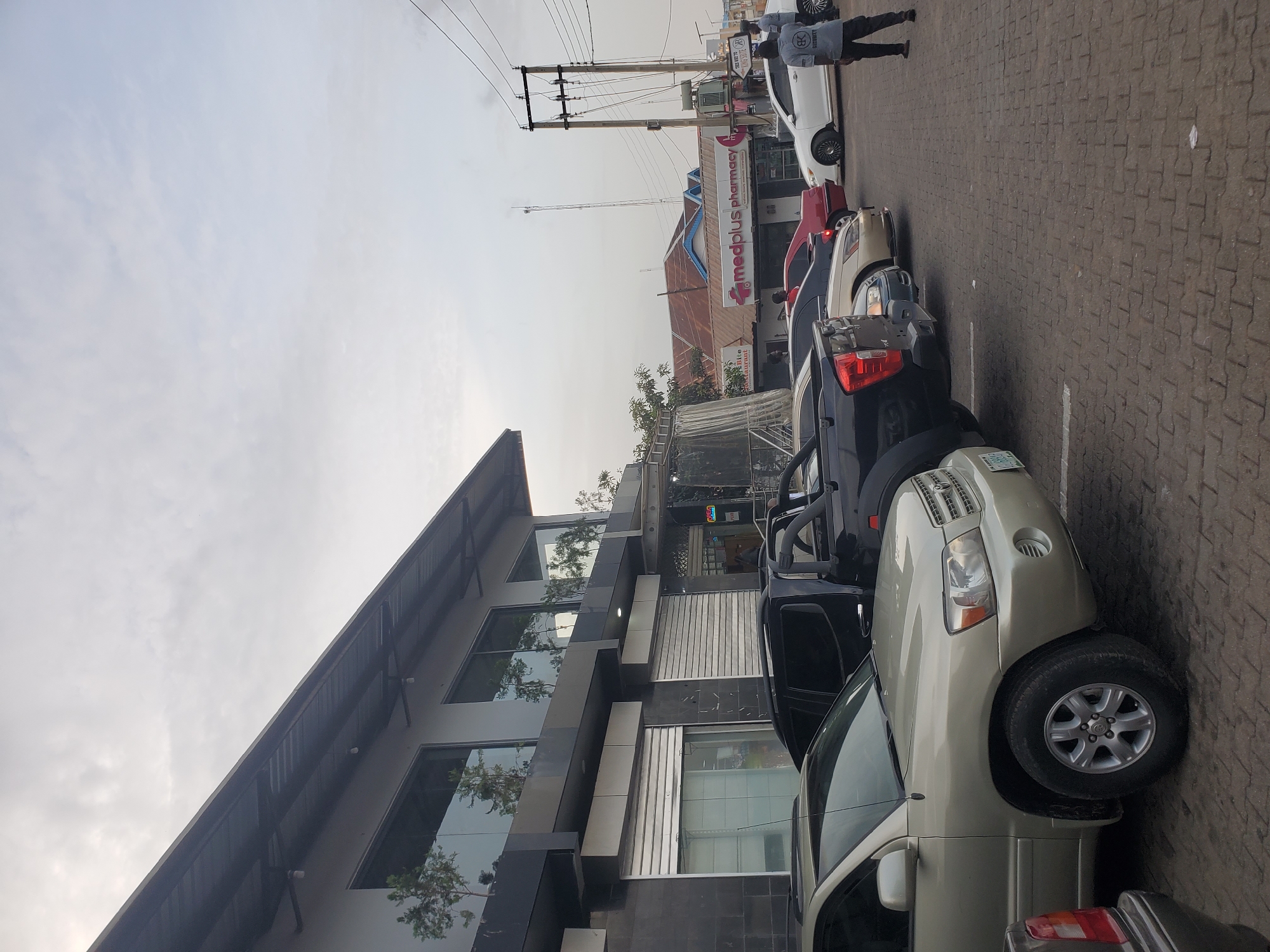 The image captures a commercial area in Jos, Nigeria, likely a shopping complex or a row of shops. In the foreground, several parked vehicles are visible, including a light-colored SUV, a black pickup truck, and other cars. The buildings feature modern architecture with large glass windows on the upper level and shuttered storefronts on the ground floor. Signs indicate the presence of businesses, with "medplus pharmacy" being clearly legible on one building. The street is paved with interlocking bricks, and utility poles with electrical wires are present. A few individuals are seen walking in the background, some near the pharmacy. The sky is overcast, suggesting a cloudy day. The overall scene depicts a typical urban commercial street with parked vehicles and pedestrian activity.