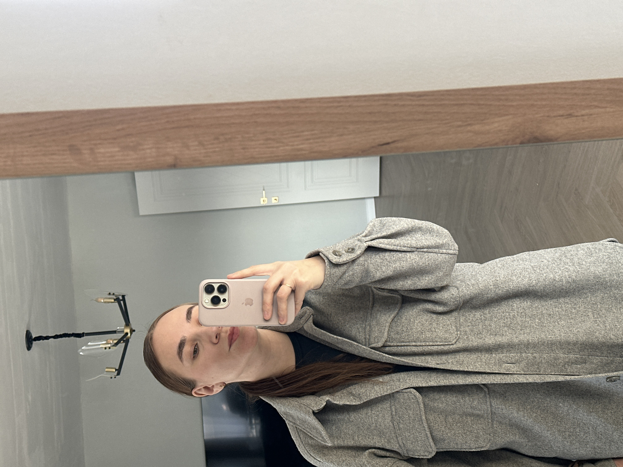A woman is taking a selfie in a mirror. She is holding a pink smartphone, which obscures part of her face. She is wearing a gray wool coat over a black shirt. The reflection shows a modern chandelier on the wall, a gray wall, a white door with gold-colored hardware, and possibly a television screen. The location is unknown, and there is no verifiable information to indicate it is Kharnavy, Russia.