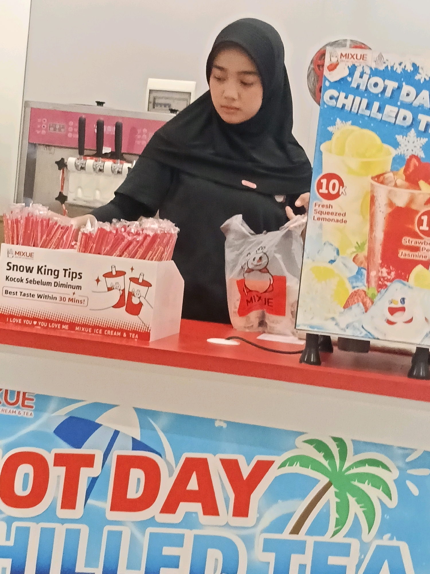 A young woman wearing a black hijab and black shirt stands behind a counter in what appears to be a convenience store or ice cream shop in Belimbing, Indonesia. She is looking down and slightly to her left, with her right hand extended towards a pink ice cream machine with three dispensers. In front of her, on the counter, are boxes of red and white striped straws labeled "MIXUE Snow King Tips". To her right, a large sign displays promotional text and images for "HOT DAY CHILLED TEA" and "Fresh Squeezed Lemonade". The sign features images of drinks and cartoon characters. A clear plastic bag with a snowman logo and "MIXUE" printed on it is placed on the counter to the left of the sign. The counter itself is red. The background shows a white wall and the ice cream machine.