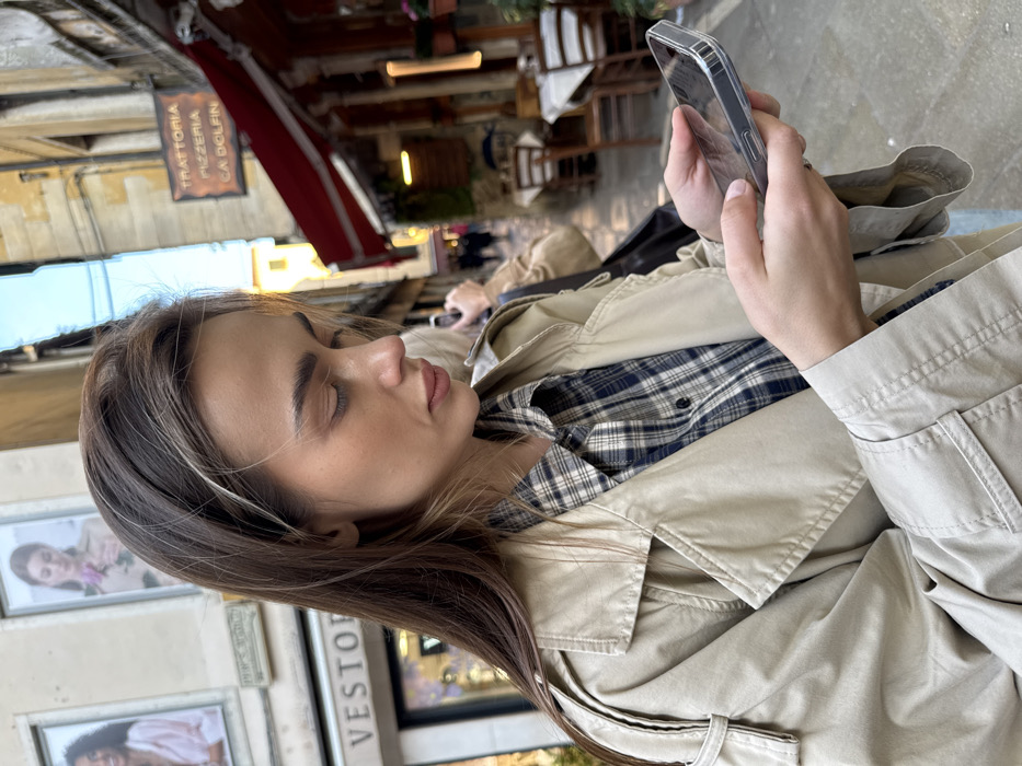 A medium close-up photograph depicts a young woman intently viewing a smartphone, positioned in a narrow street in Venice, Italy. The woman, with long, dark hair featuring light brown highlights, wears a beige trench coat over a dark blue and white plaid shirt. Her eyes are downcast, focused on the screen of her clear-cased smartphone, held with both hands.
To the left of the woman, a building features display windows containing fashion advertisements, including one depicting a woman holding a pink rose and another of a woman in a pink top. A partial sign "VESTOR" is visible below the windows.
To the right and in the background, a traditional Venetian calle (alleyway) recedes. A prominent sign "TRATTORIA PIZZERIA CA DOLFIN" is affixed to a building with a red awning. Beneath the awning, outdoor dining tables draped with white tablecloths and accompanied by wooden chairs are arranged. Blurred figures of individuals are discernible further down the calle. Another person, mostly out of focus, stands behind the primary subject, also seemingly engaged with a mobile device. The street is paved with traditional stones, and a strip of clear blue sky is visible above the buildings. The location is identified as Calle Larga Ca' Dolfin, 4821, 30123 Venezia VE, Italy, based on the trattoria sign.