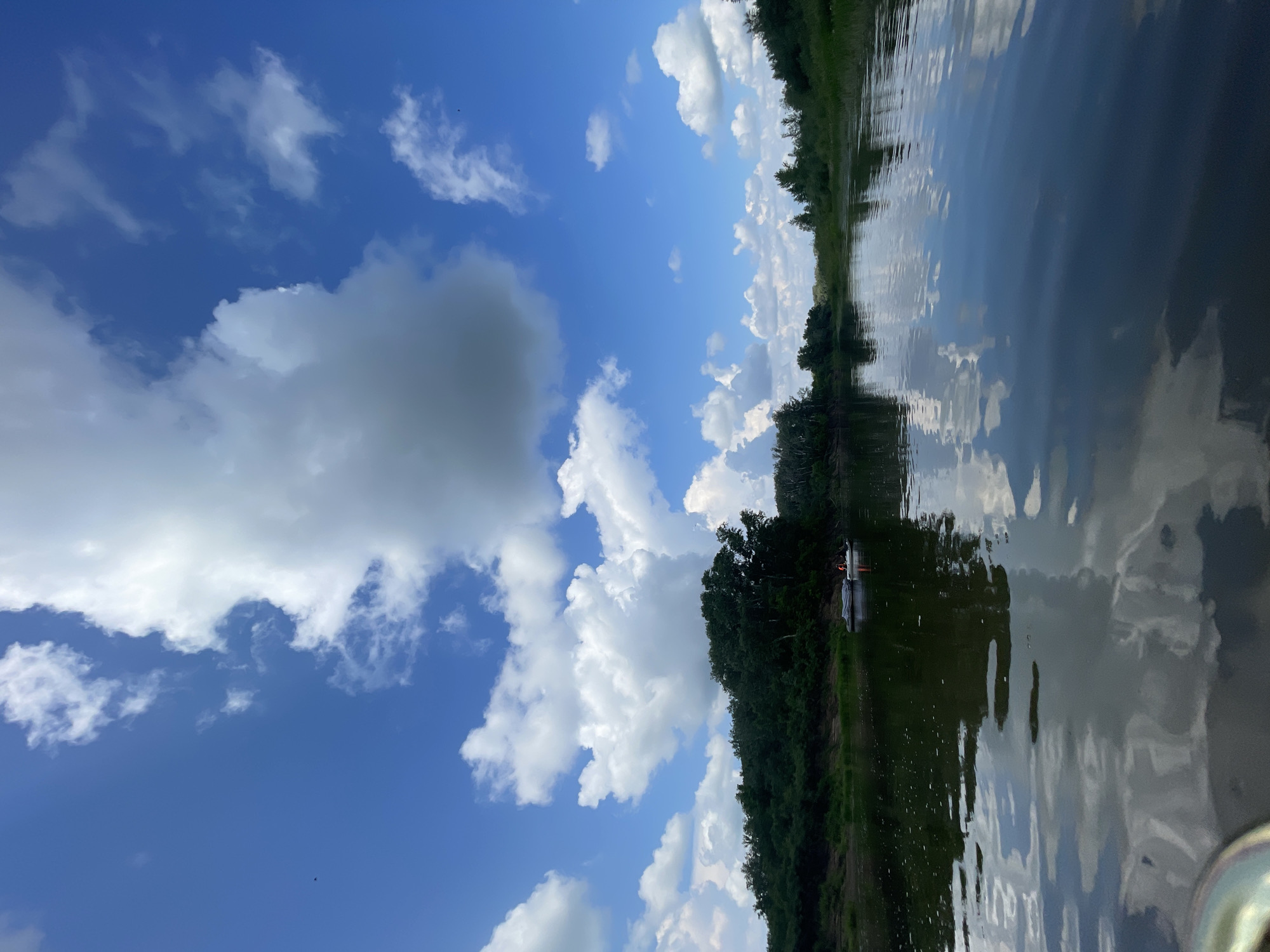 A wide, calm river flows through a lush green landscape under a vibrant blue sky filled with large, puffy white clouds. The river's surface reflects the sky and the surrounding trees, creating a mirror-like effect. On the river, a small boat with two people wearing orange life vests is visible near the left bank, which is lined with dense green foliage and trees. The scene appears peaceful and natural, likely a popular spot for outdoor recreation.