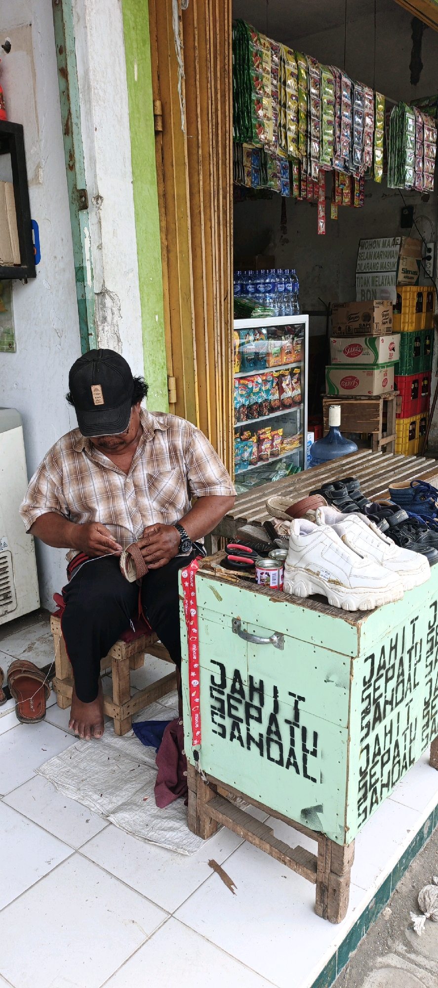 In Kasiyan, Indonesia, a man wearing a black baseball cap and a plaid shirt is seated on a wooden stool, diligently sewing a brown sandal. His black pants are tucked into his bare feet, which rest on a white tarp laid out on the tiled floor. To his right, a light green wooden box with the words "JAHIT SEPATU SANDAL" stenciled in black stands on wooden legs. Several pairs of shoes and sandals are displayed on a slatted wooden surface above the box, including a pair of white chunky sneakers. The background reveals a shop entrance with a yellow roll-up door, shelves stocked with various goods, including bottled water and packaged snacks, and stacks of colorful crates. The scene captures a street-side shoe repair service.