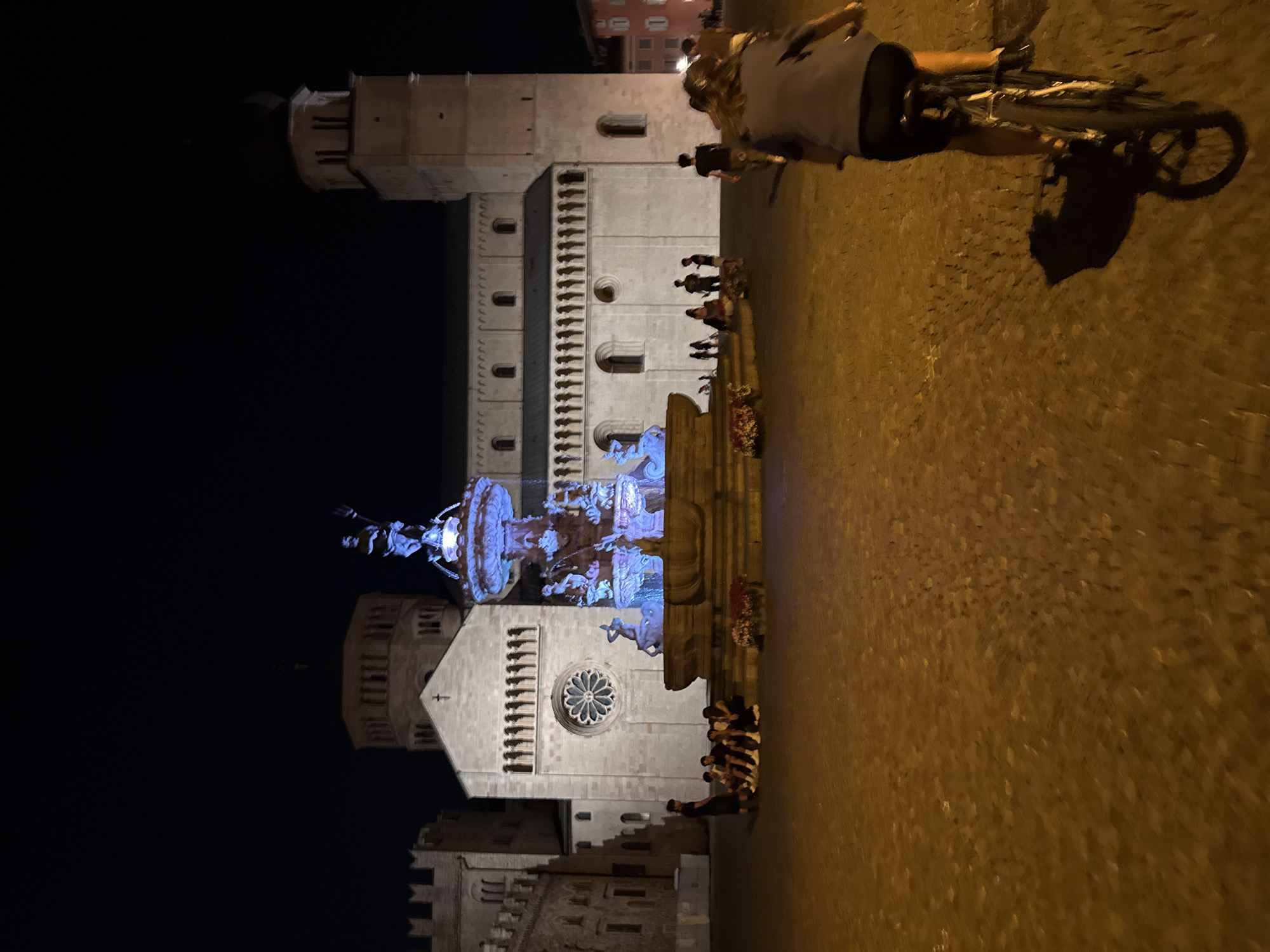 This is a nighttime, low-angle shot of Piazza Duomo in Trento, Italy. A group of people are gathered in front of a large fountain adorned with blue lights. A woman is riding a bicycle in the foreground, casting a shadow on the cobblestone ground. Behind the fountain, the Duomo of Trento is visible, illuminated by artificial lights. Other buildings and a tower are also present. The scene is dark, with artificial lighting creating illuminated areas and deep shadows.