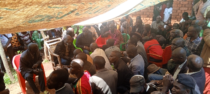 A large group of people are gathered under a tent-like structure, with many seated in plastic chairs. The attendees are diverse in age and attire, some wearing casual clothing, others in suits or traditional garb. The setting appears to be outdoors, possibly a community event or gathering. Brickwork is visible in the background behind the crowd. The lighting suggests it is daytime.