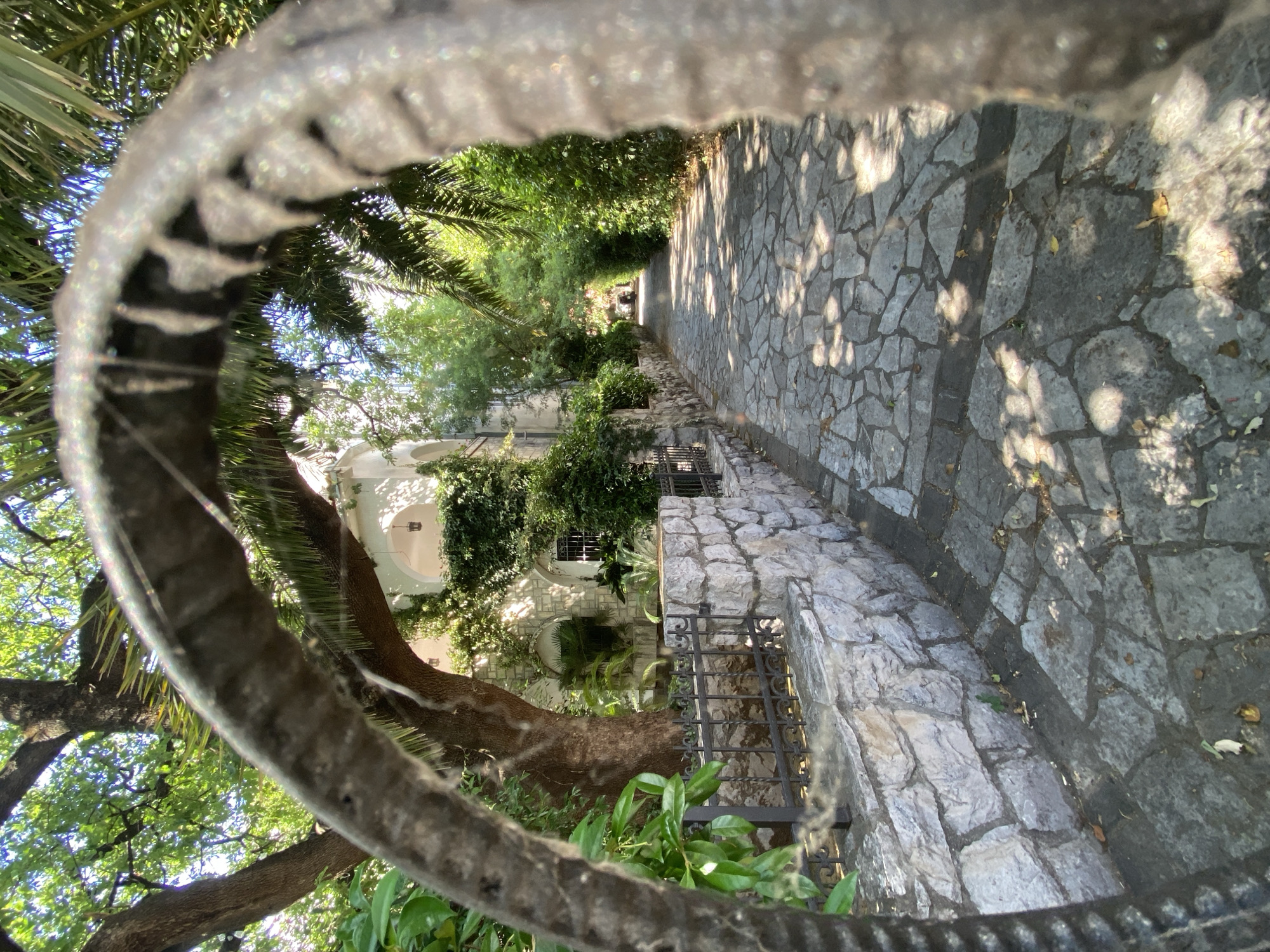 The image captures a sun-dappled stone pathway bordered by a low stone wall and lush greenery, leading towards a building with arched entrances and white walls. The pathway is paved with irregularly shaped stones and casts shadows from the surrounding trees. A dark, rough-textured object, possibly a piece of rebar covered in dust or fibers, is prominently in the foreground, framing the view. The scene appears to be a tranquil residential area, likely in the city of Budva, Montenegro, given the architectural style and Mediterranean vegetation. No people are visible in the image.