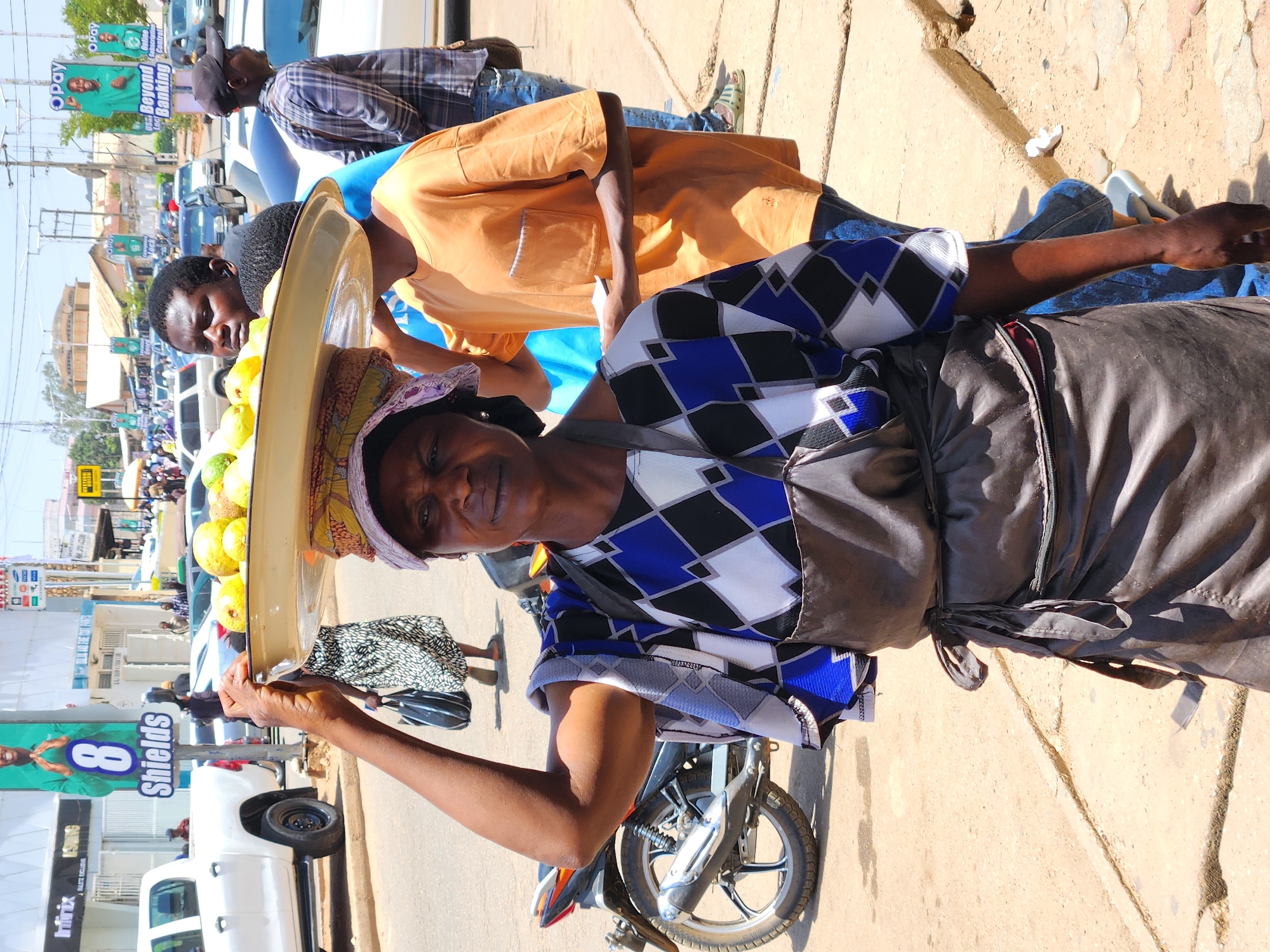 In an outdoor scene of an unknown city, an African woman stands on a sidewalk, balancing a yellow tray of round, yellowish-green fruits on her head. She's wearing a colorful top with geometric patterns of white, black, and blue, paired with a gray skirt or wrap. A motorcycle is parked near her, and she holds the edge of the tray with her left hand.
Behind her, a man in an orange shirt stands on the sidewalk, along with others in the background. Cars and various signs are visible in the street behind them, including a sign with the word "Shields" on it. The sunlight casts strong shadows on the ground, and the overall scene appears to be a bustling street market.