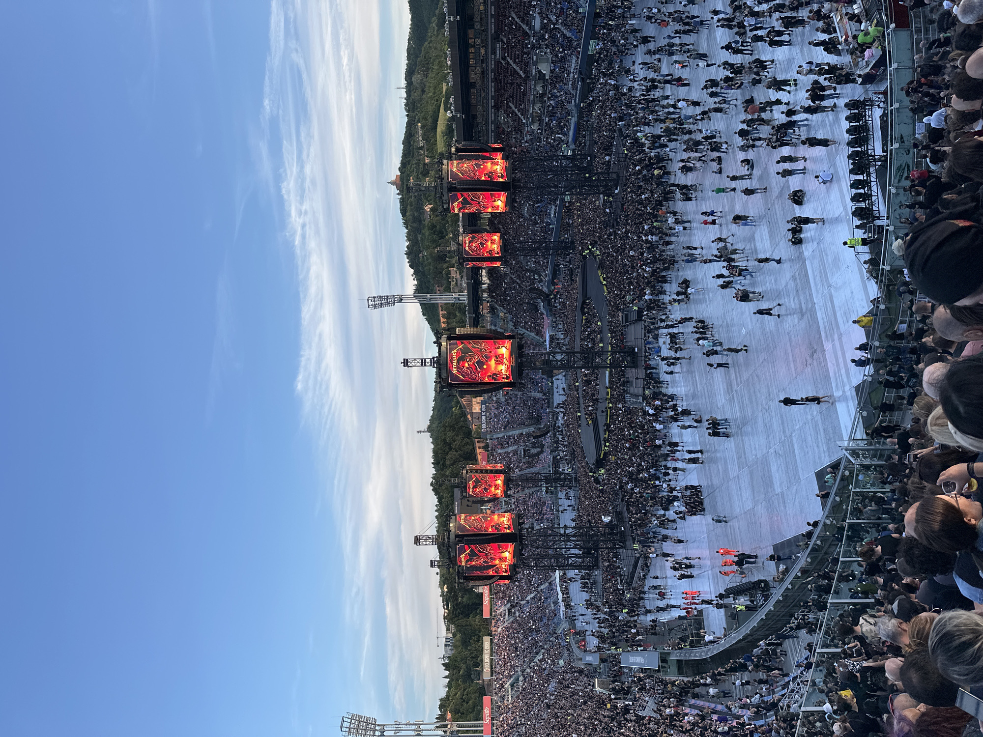 This image captures a large-scale outdoor music concert or festival, viewed from an elevated position within the stadium stands, likely during late afternoon or early evening. Thousands of people fill the venue, with a vast crowd spread across the field in front of the stage and others occupying the stadium seating.
At the heart of the scene is an impressive stage setup featuring multiple towering LED video screens. These screens display vibrant red and black graphics, and the band's name "METALLICA" is clearly visible on at least one, identifying the event as a Metallica concert. The screens are integrated into large support structures that also hold lighting and sound equipment.
The sky is a clear blue with wispy white clouds, indicating pleasant weather. In the background, beyond the stadium, tree-covered hills are visible, with a prominent historical building or castle situated on one of the hilltops, serving as a distinctive landmark. This landscape, combined with the specified location context, suggests the venue is likely the Renato Dall'Ara Stadium in Bologna, Italy.
Attendees are gathered in large numbers, mostly standing on the light-colored field area directly in front of the stage. The foreground shows the backs of audience members, indicating the photograph was taken from within the crowd. Along the perimeter of the field, several white banners are visible, displaying text such as "SAPORITO," "MERCATO," "PROSECCO DOC," and "GIUSTO," likely sponsors of the event. The scene conveys the excitement and anticipation typical of a major music performance.